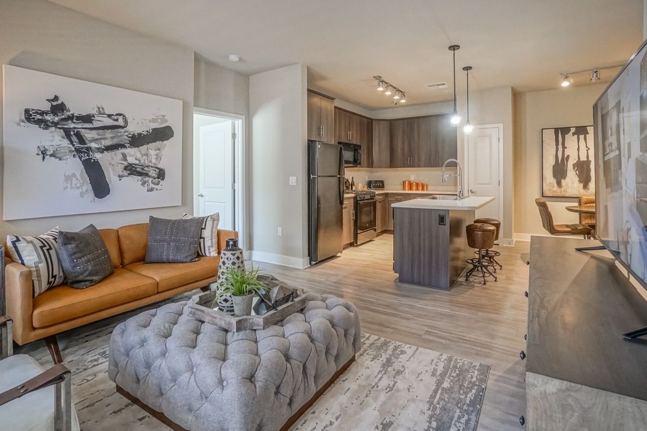 Open-concept living area with a tan sofa, tufted gray ottoman, and a modern kitchen island.