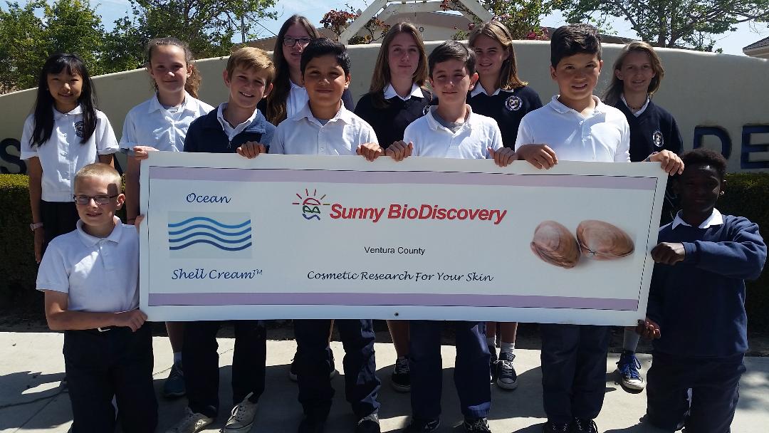 Children holding an oversized check outdoors, near a sign that says