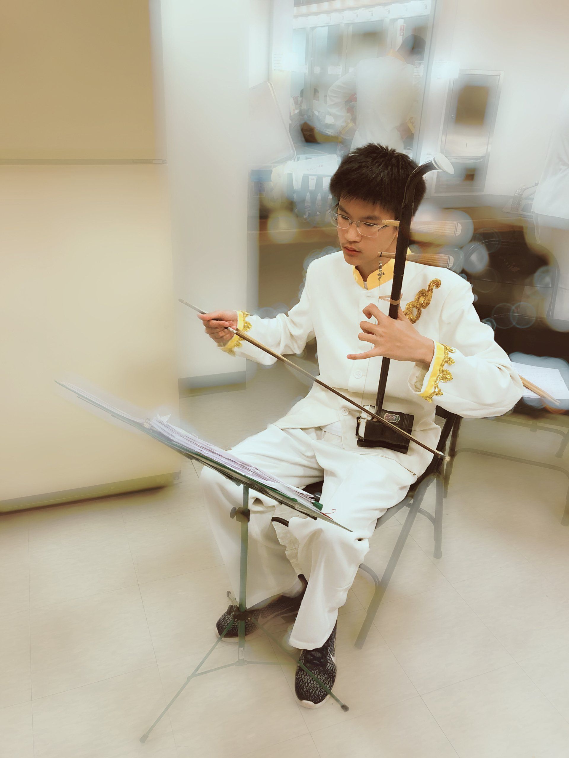 Person in white outfit playing an erhu, a Chinese two-stringed bowed instrument. Seated, sheet music in front.