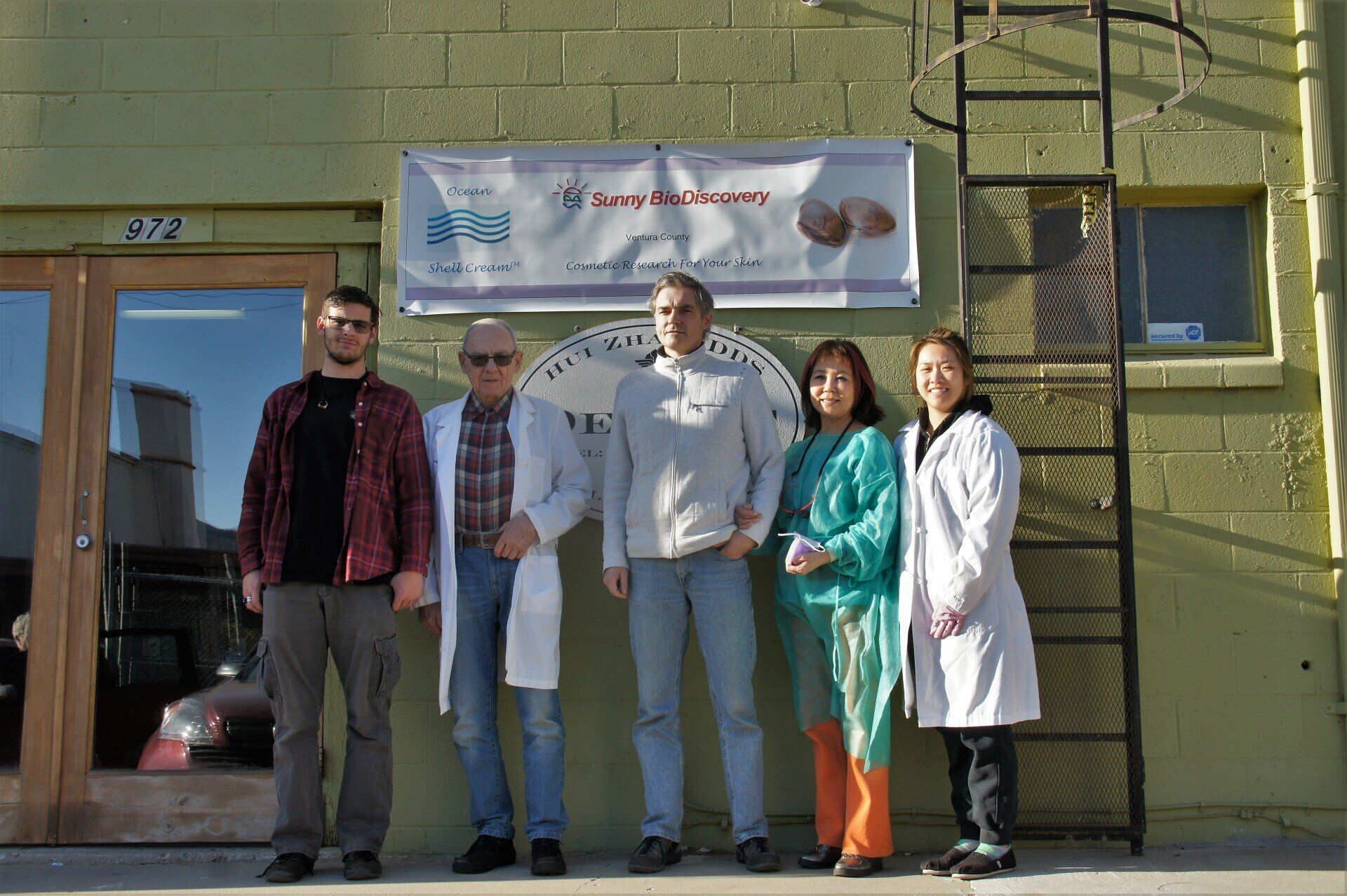 Group of five people in front of a building with a sign. Sunny day, two wear lab coats.