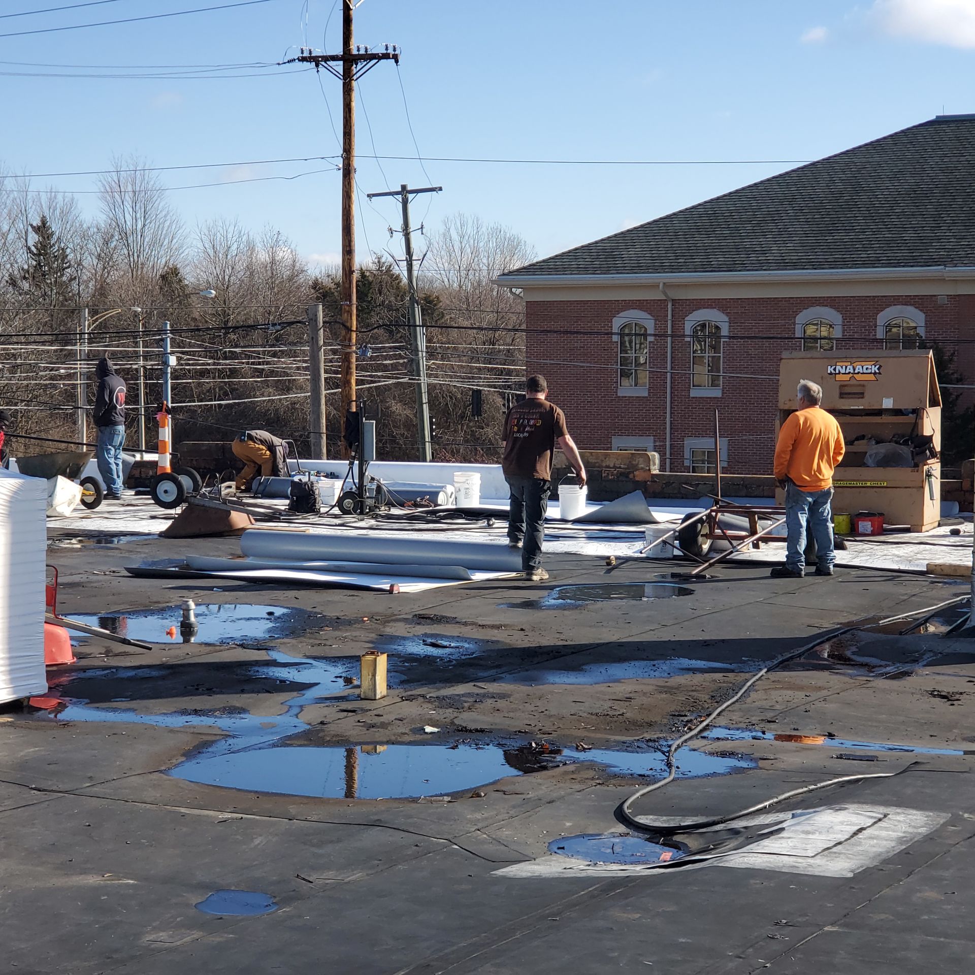 Workers on a construction site outside a brick building. Puddles and debris on asphalt; clear sky.