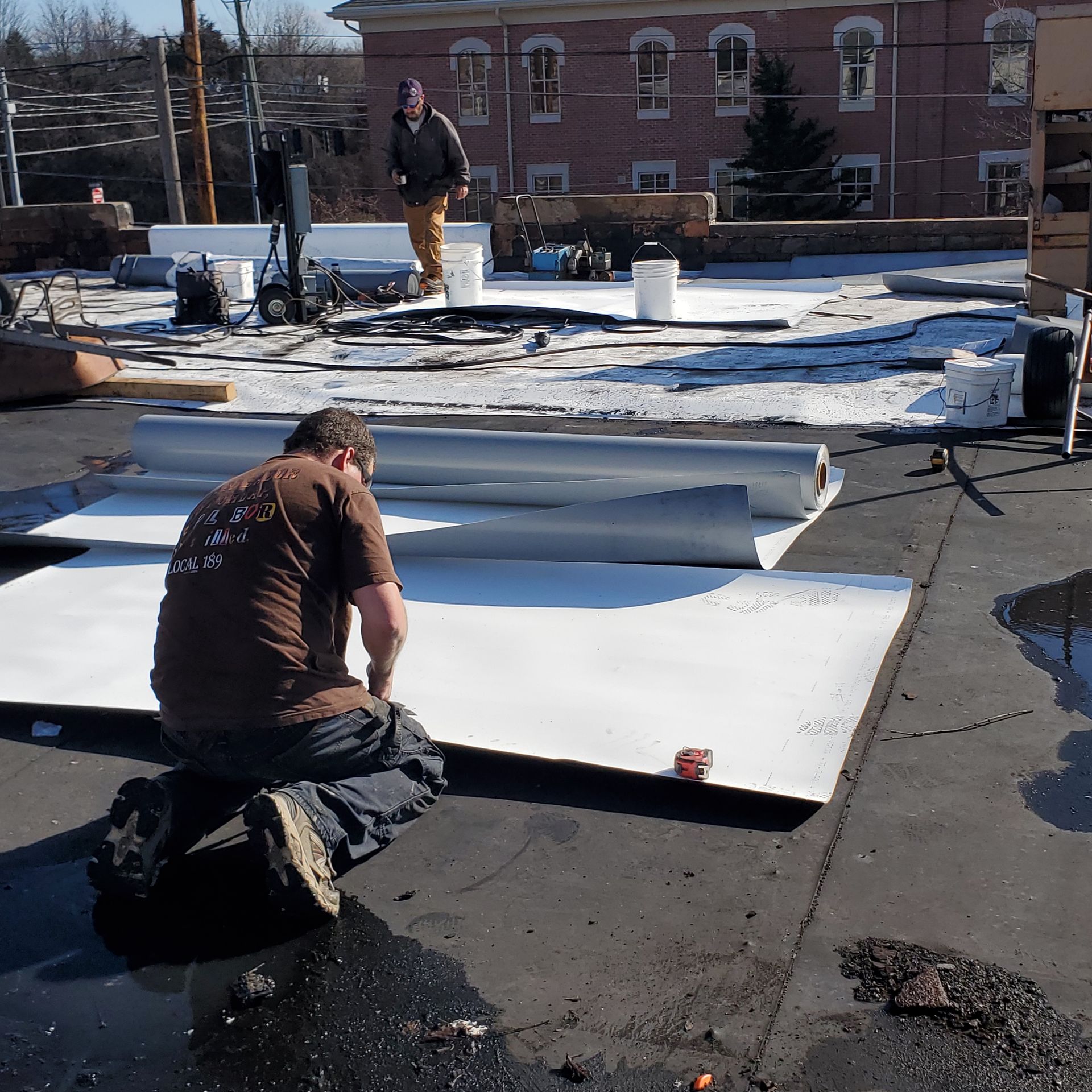 Construction workers on a flat roof. One kneels, looking at a large white sheet. Another stands in the background.