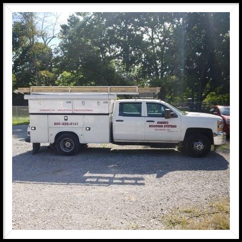 White work truck with a storage bed; parked on gravel; surrounded by trees.
