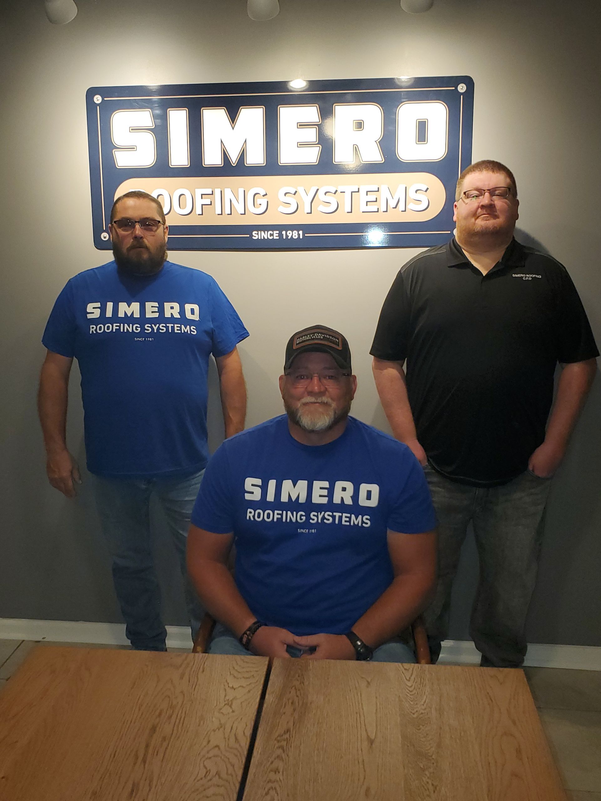 Three men in blue shirts pose in front of a Simero Roofing Systems sign.