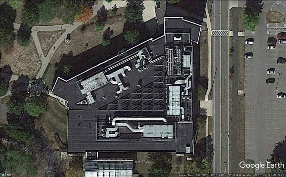 Overhead view of a modern building with a complex roof structure, surrounded by trees, parking, and a road.