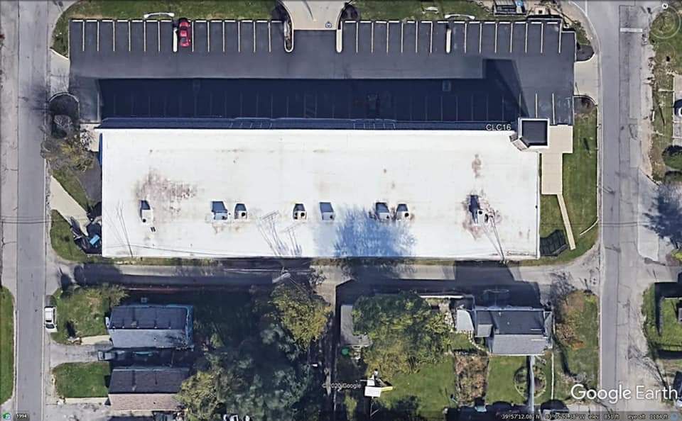 Aerial view of a long, flat-roofed commercial building with a parking lot. Street and homes are visible.