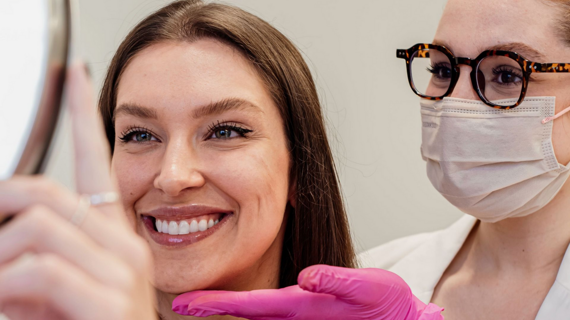Dentist wearing pink gloves examines a smiling patient’s teeth with a mirror