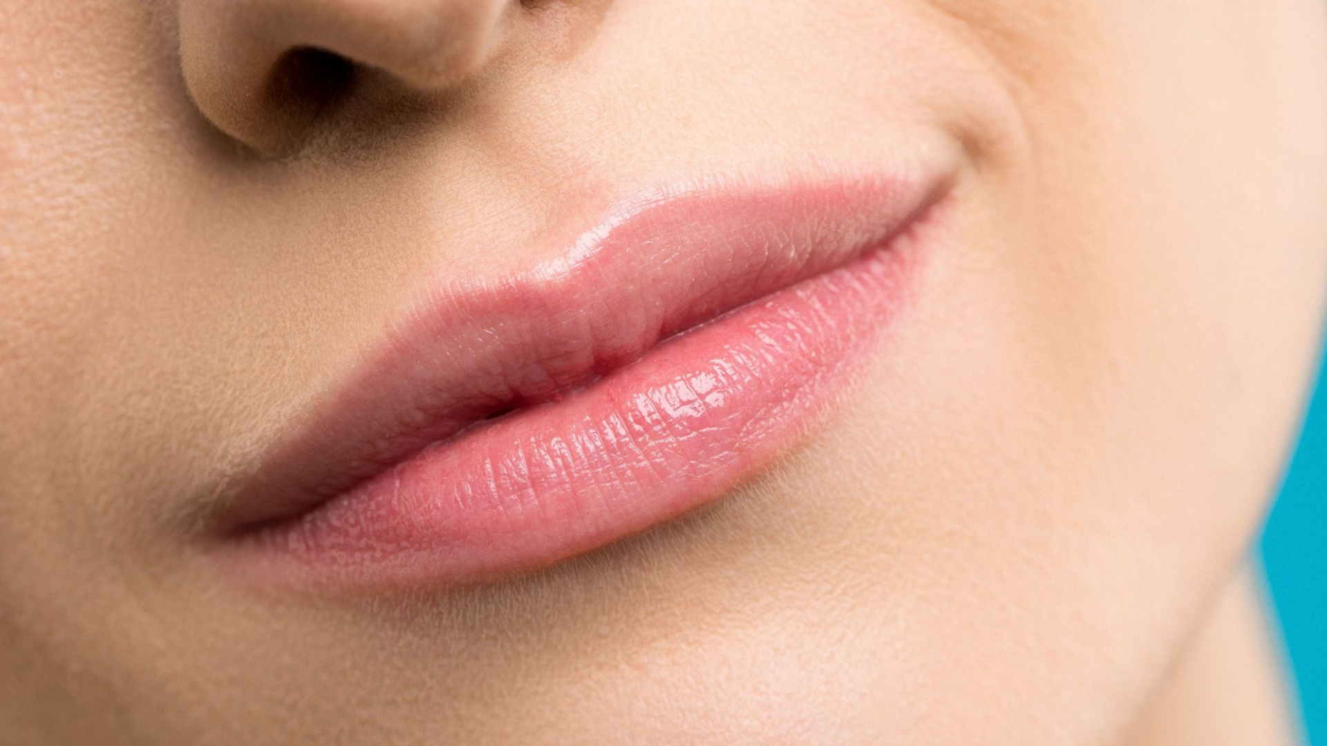 Close-up of a woman’s lower face with glossy pink lips against a blue background