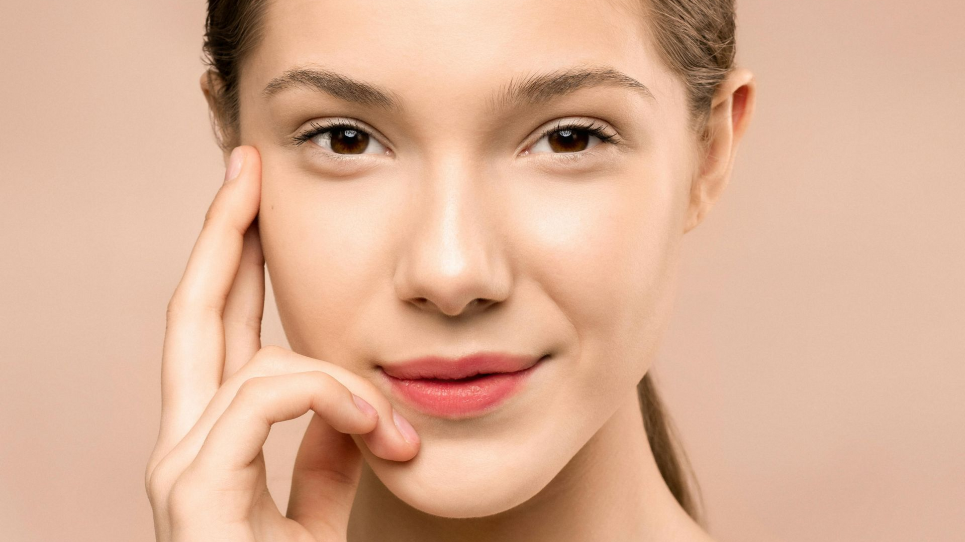 Close-up of a person touching their cheek, with a neutral expression against a soft pink background