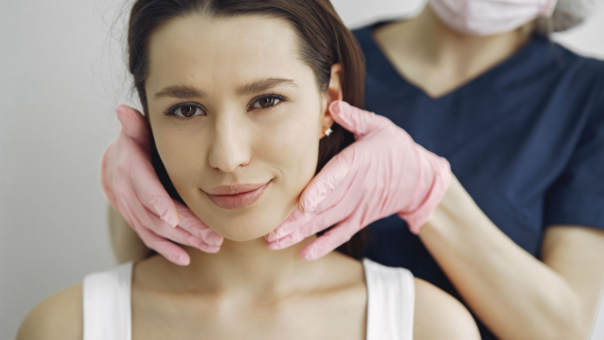 Cosmetic treatment with gloved hands gently framing a woman’s face in a clinic setting