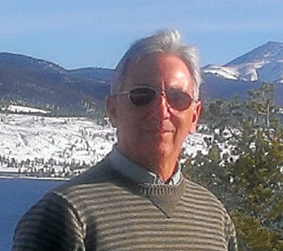 A person wearing sunglasses and a striped sweater outdoors, with a snowy mountain landscape and water in the background.