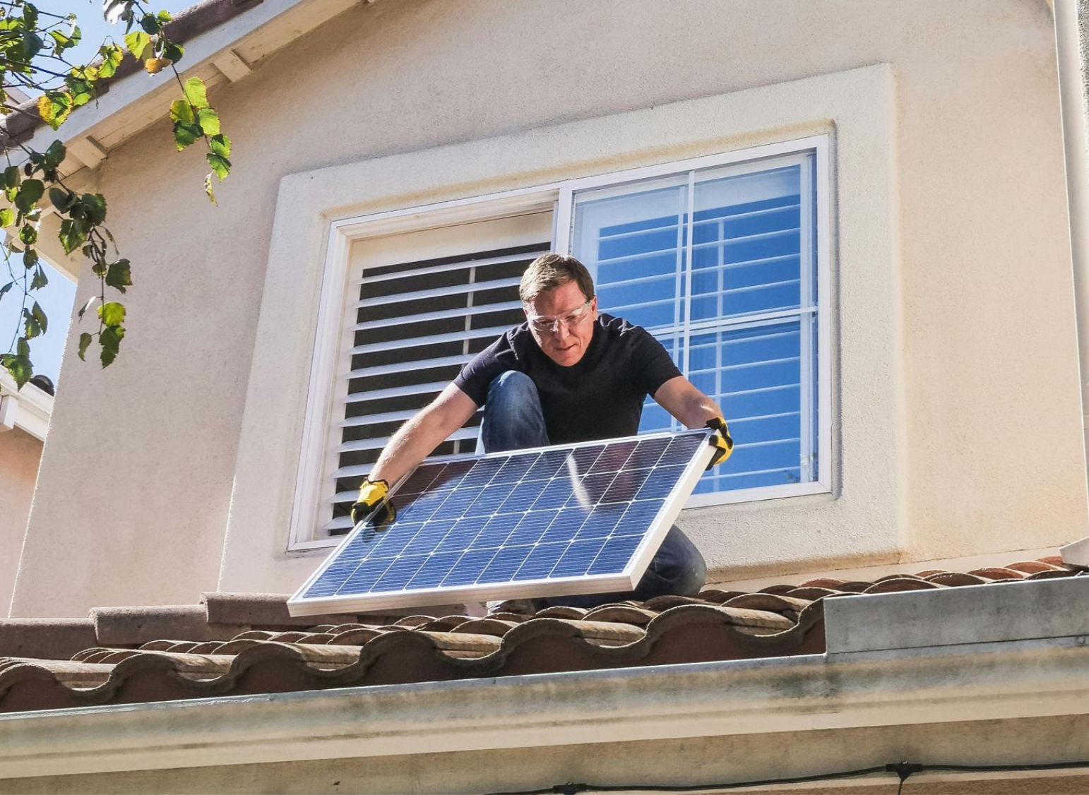 A person kneels on a residential tile roof, carefully positioning a solar panel near an open window.
