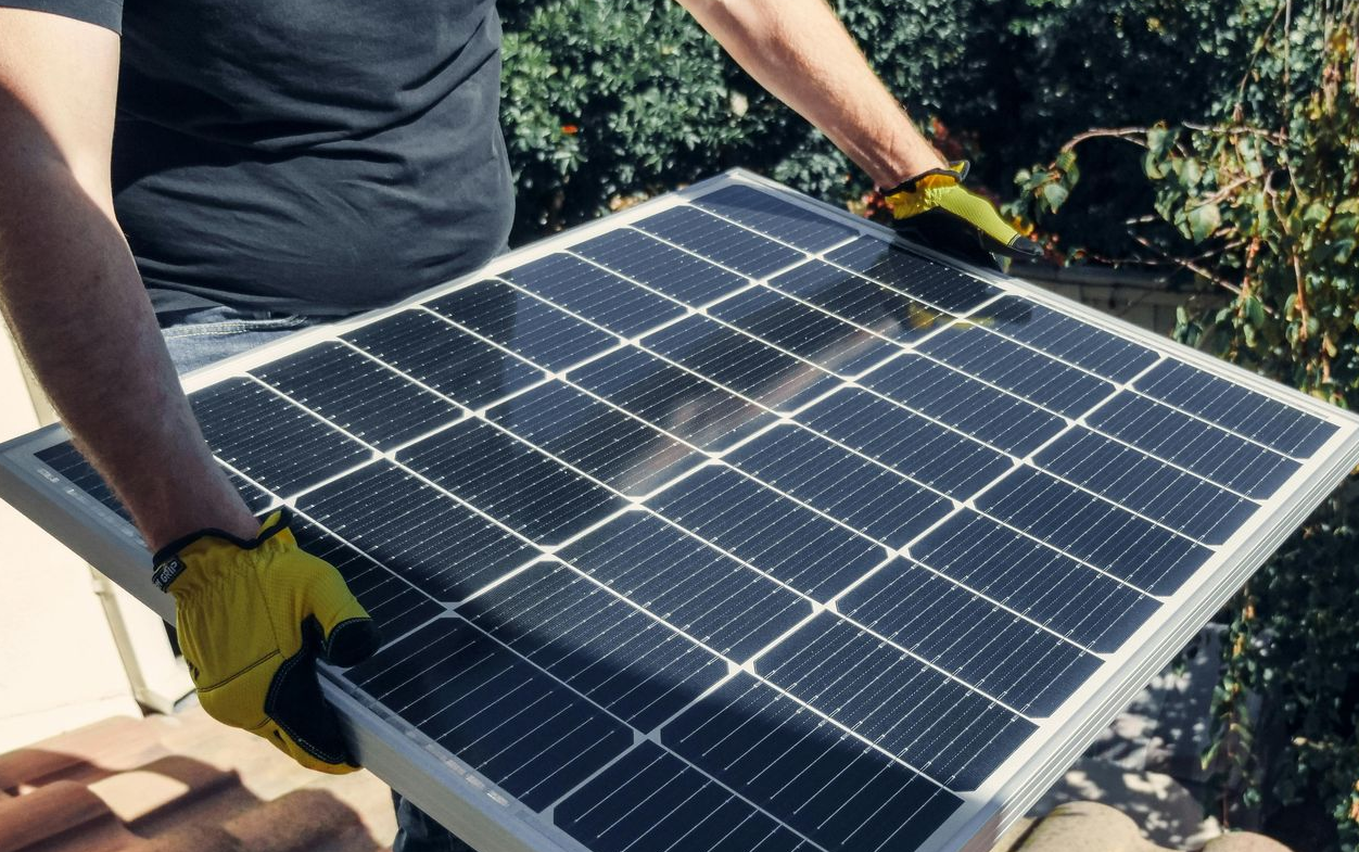 A person wearing yellow work gloves holds a rectangular solar panel outside near green foliage.