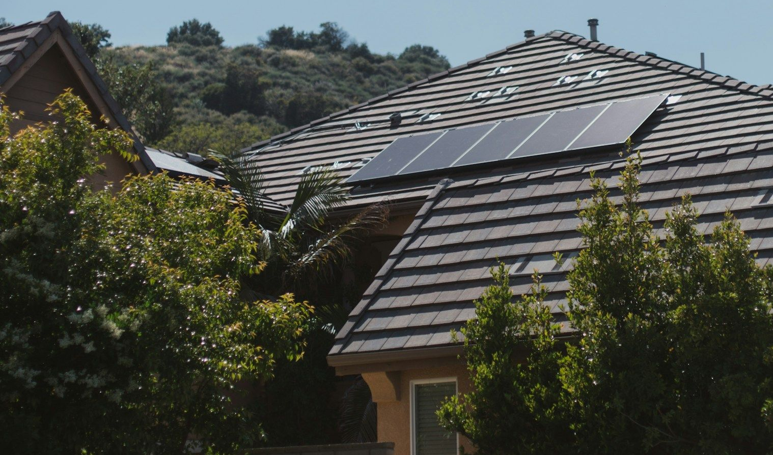 A solar panel array installed on the dark-tiled roof of a house, surrounded by lush green trees and a hillside backdrop.