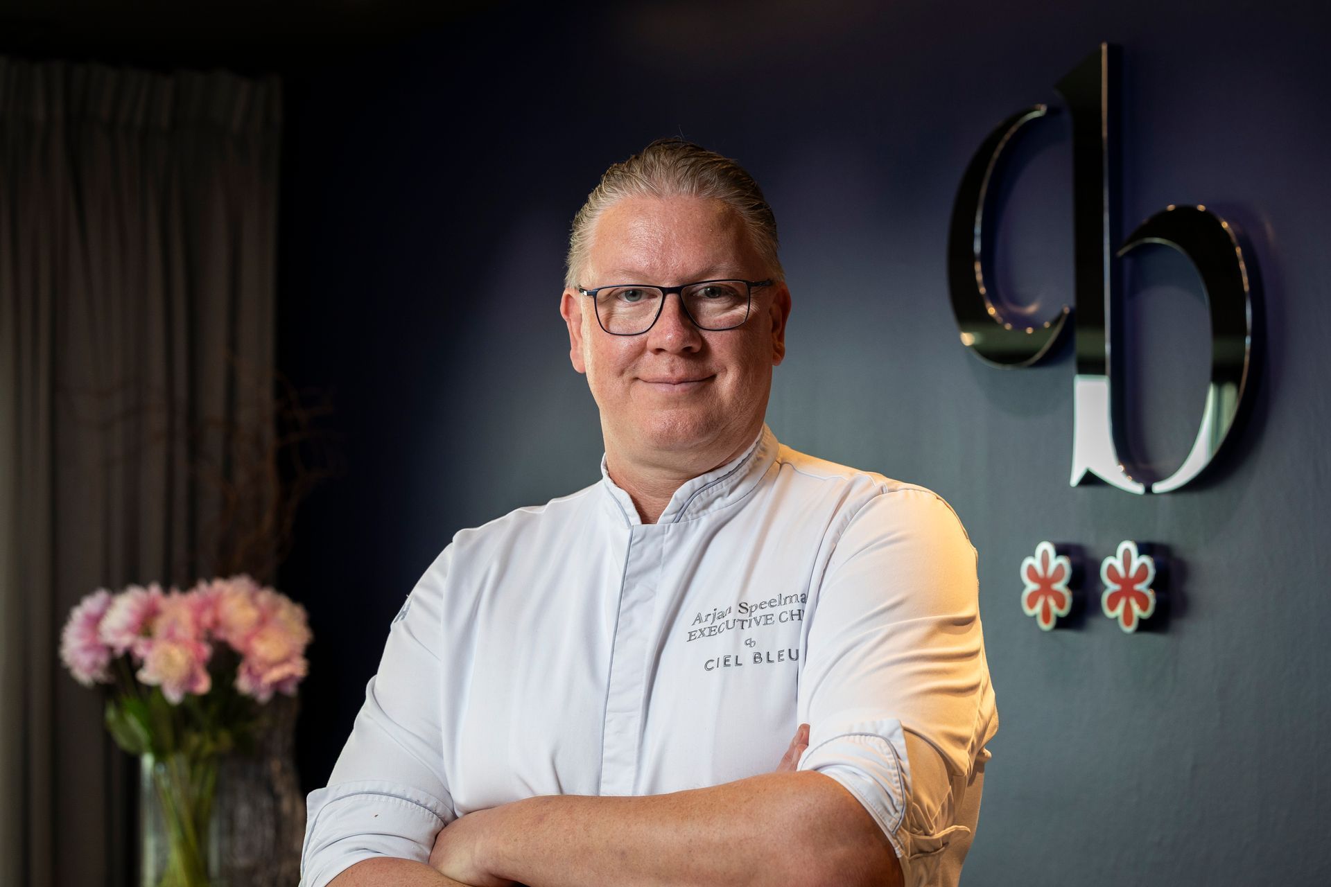 Man in white chef's coat with arms crossed, standing near a wall with a logo and flowers.