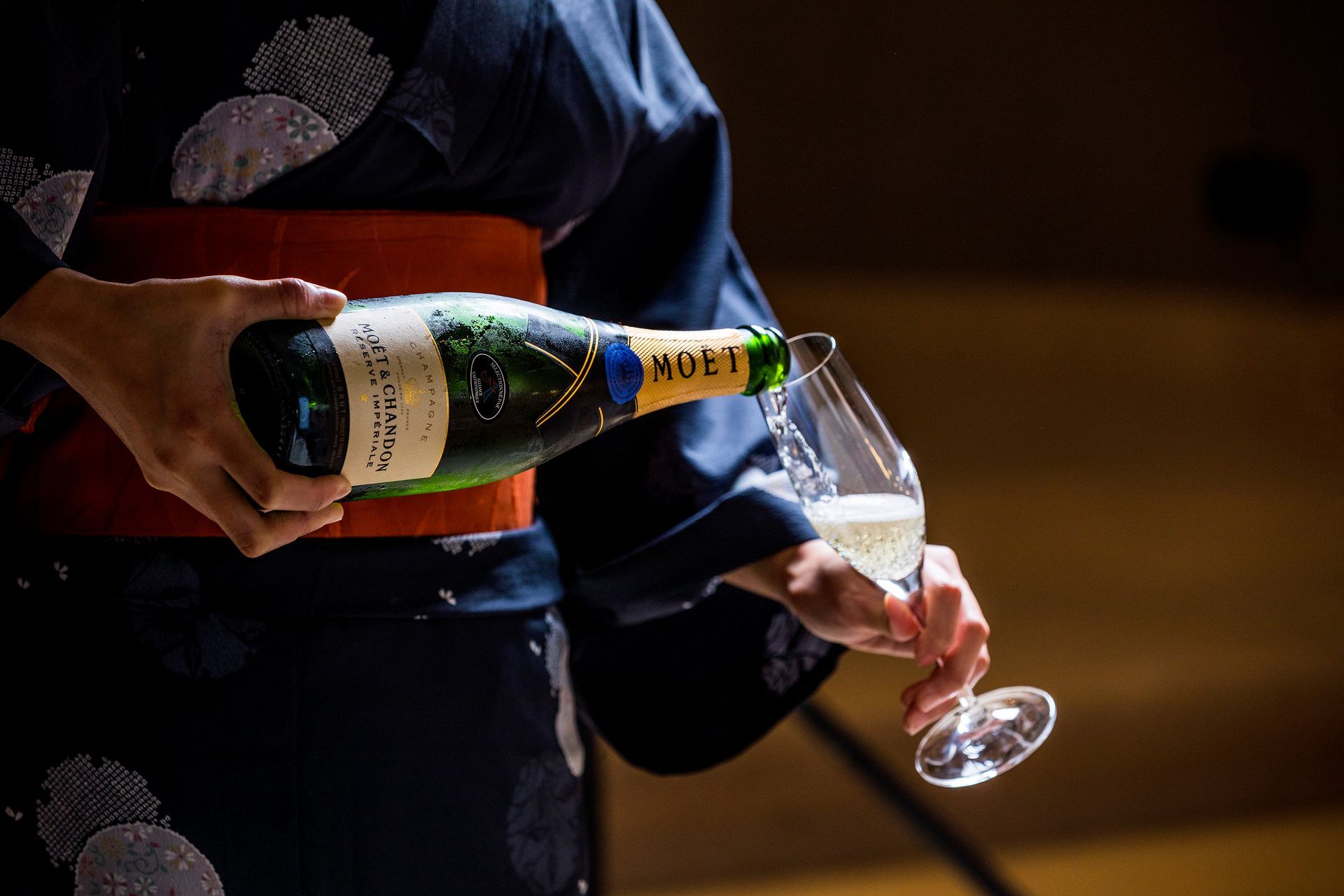 Person pouring champagne into a glass; they wear a dark blue kimono with orange sash.