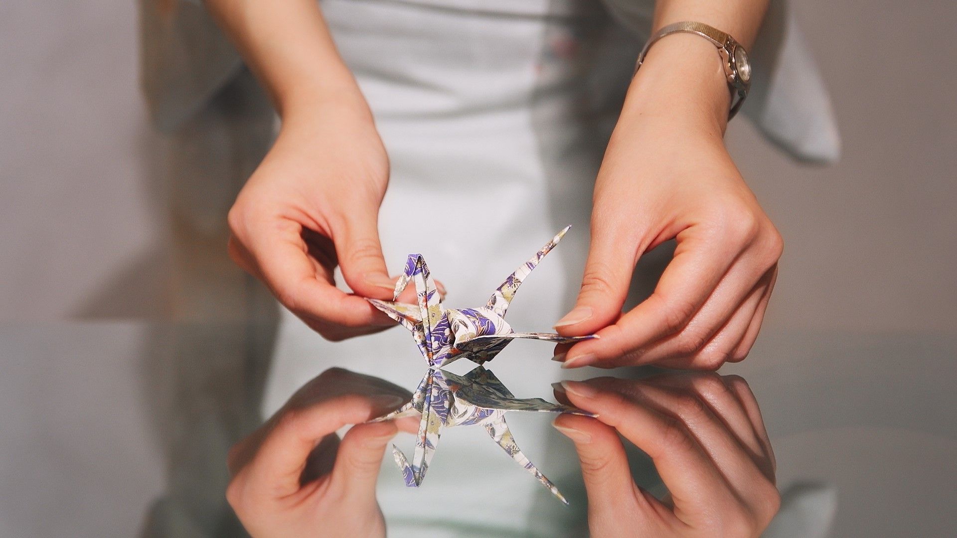 Hands holding a patterned origami crane above a reflective surface.