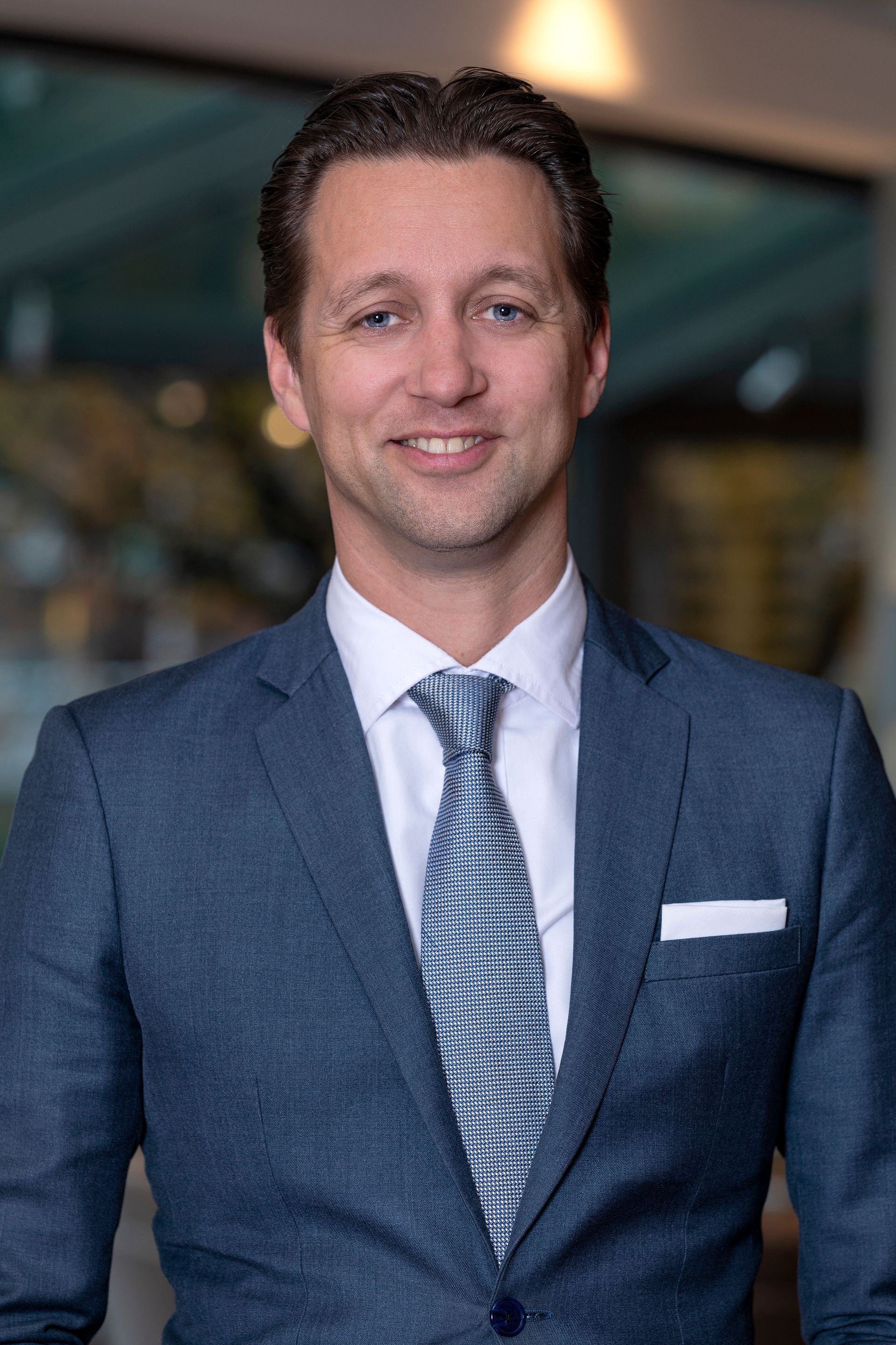 Man in a blue suit, white shirt, and patterned tie smiling in an office setting.