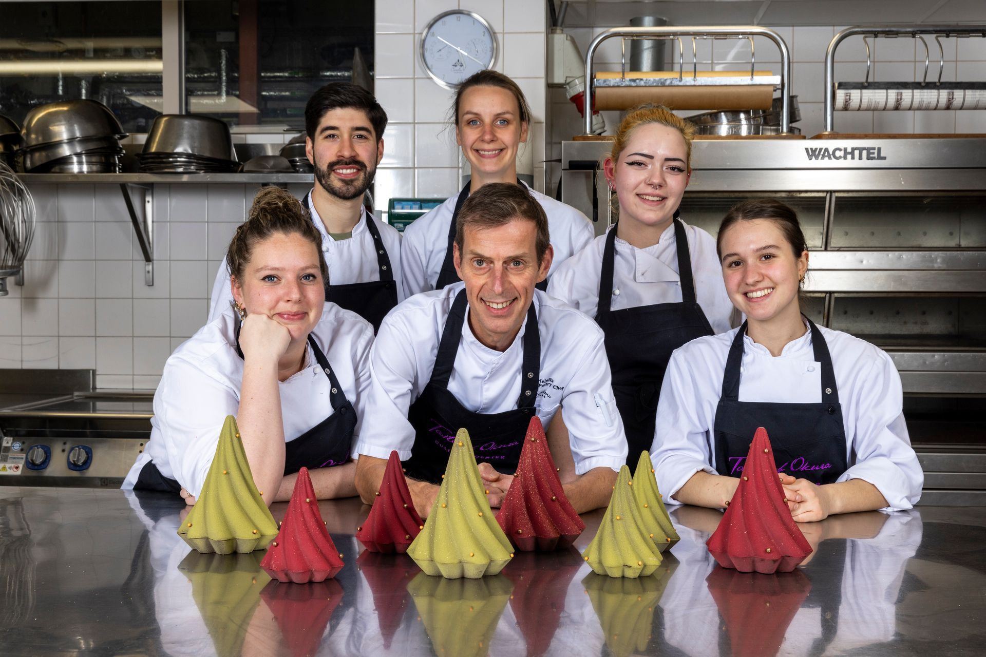 A chef and six team members pose with colorful desserts in a professional kitchen.