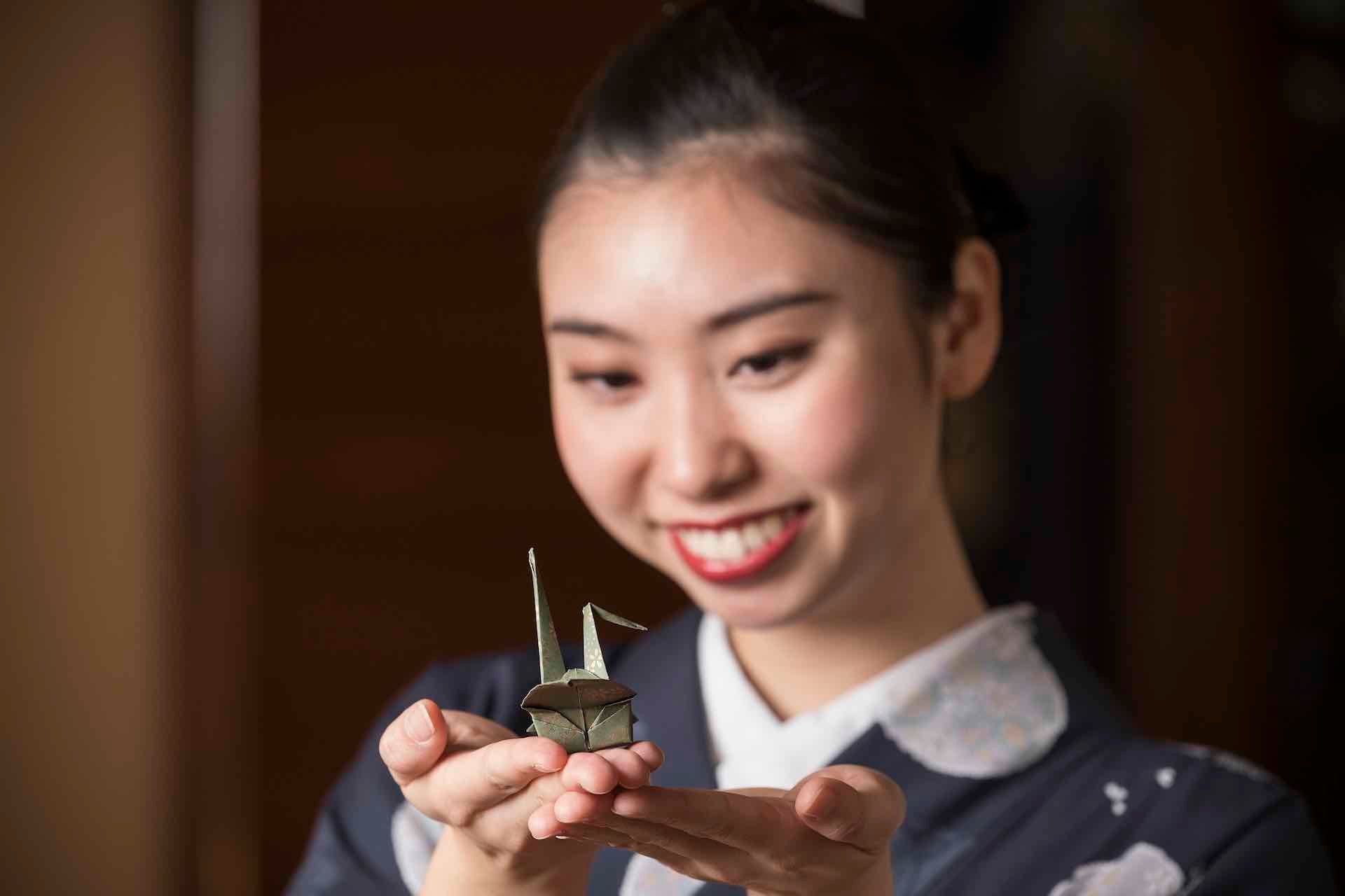 Woman in kimono smiles, holding a tiny origami crane between her hands.