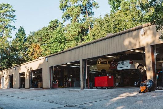 Open auto repair garage with several bays, a red tool chest, and cars inside, surrounded by trees.