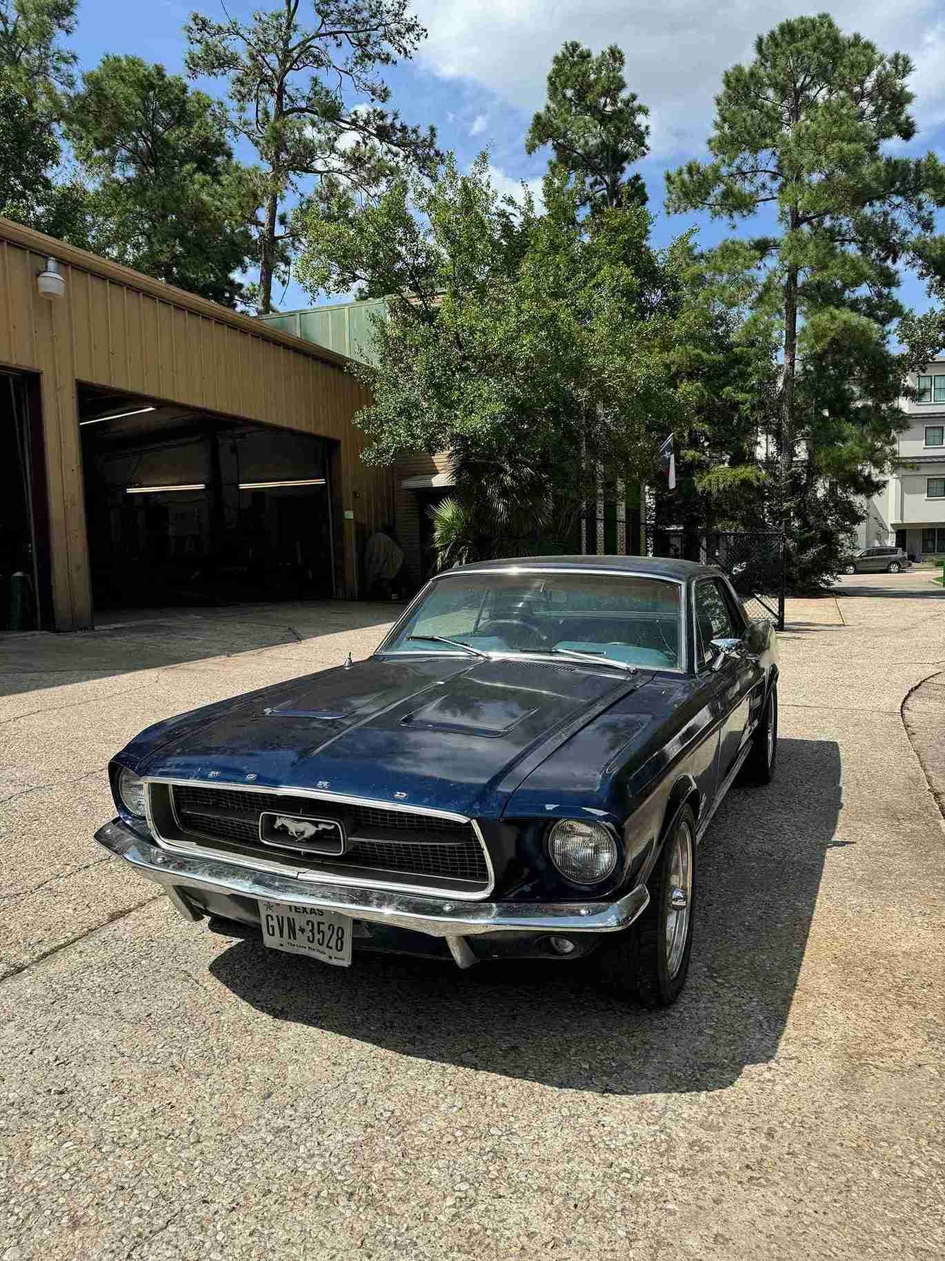 Black classic Ford Mustang parked on a gravel lot beside a garage and trees