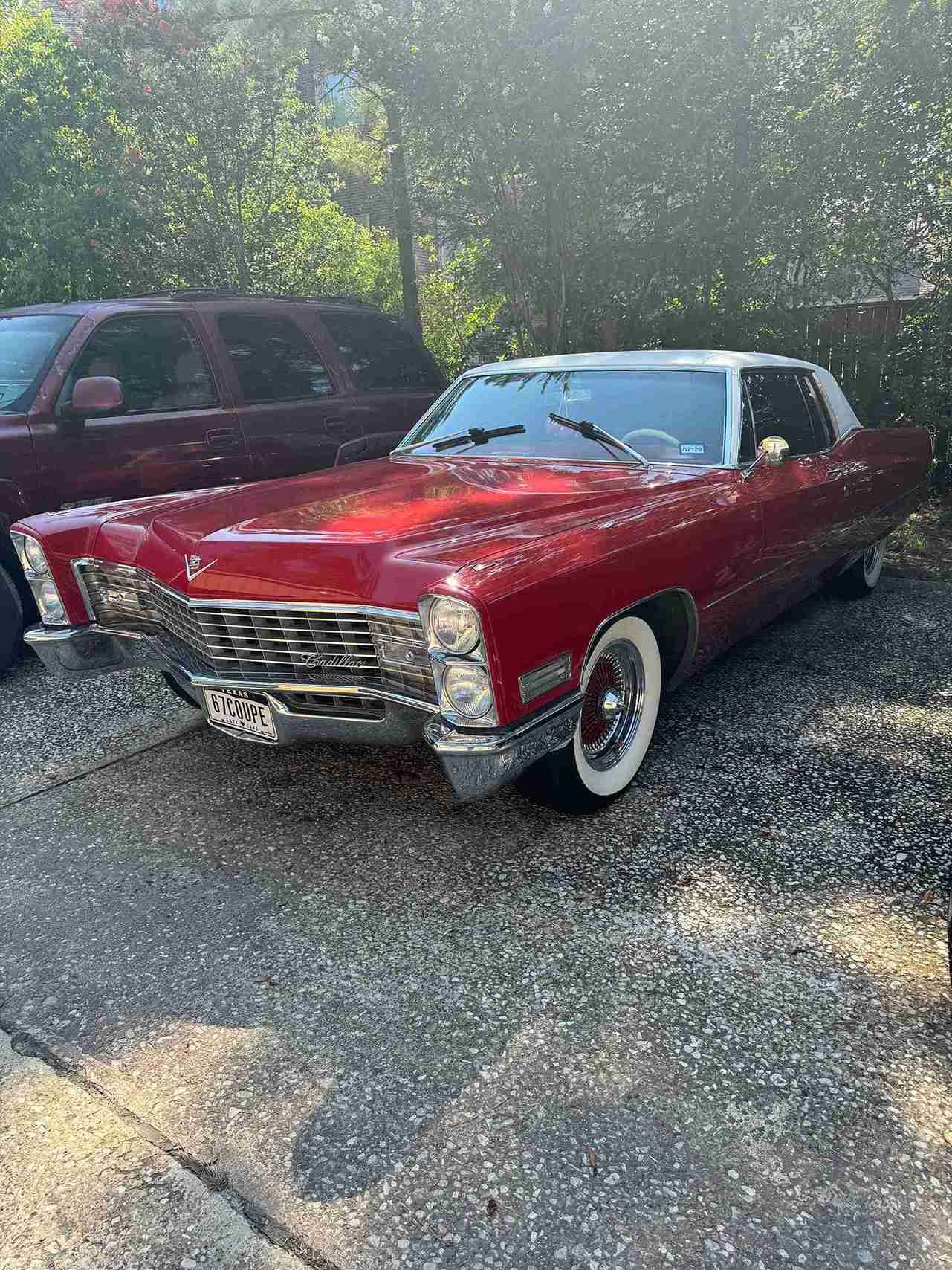 Red classic convertible parked on gravel in a wooded area, with a maroon SUV behind it.
