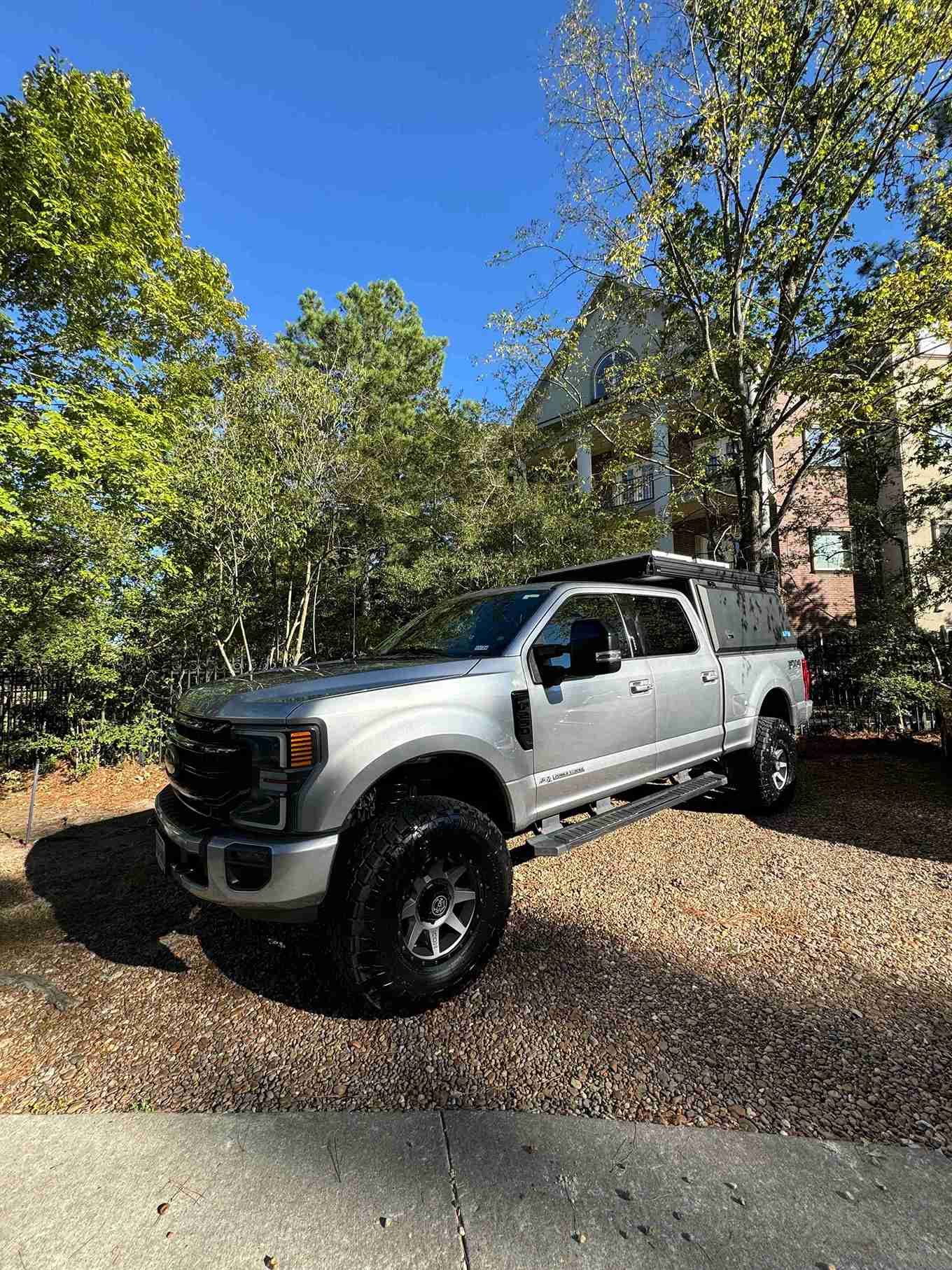 Silver pickup truck parked on a gravel driveway under trees beside a house