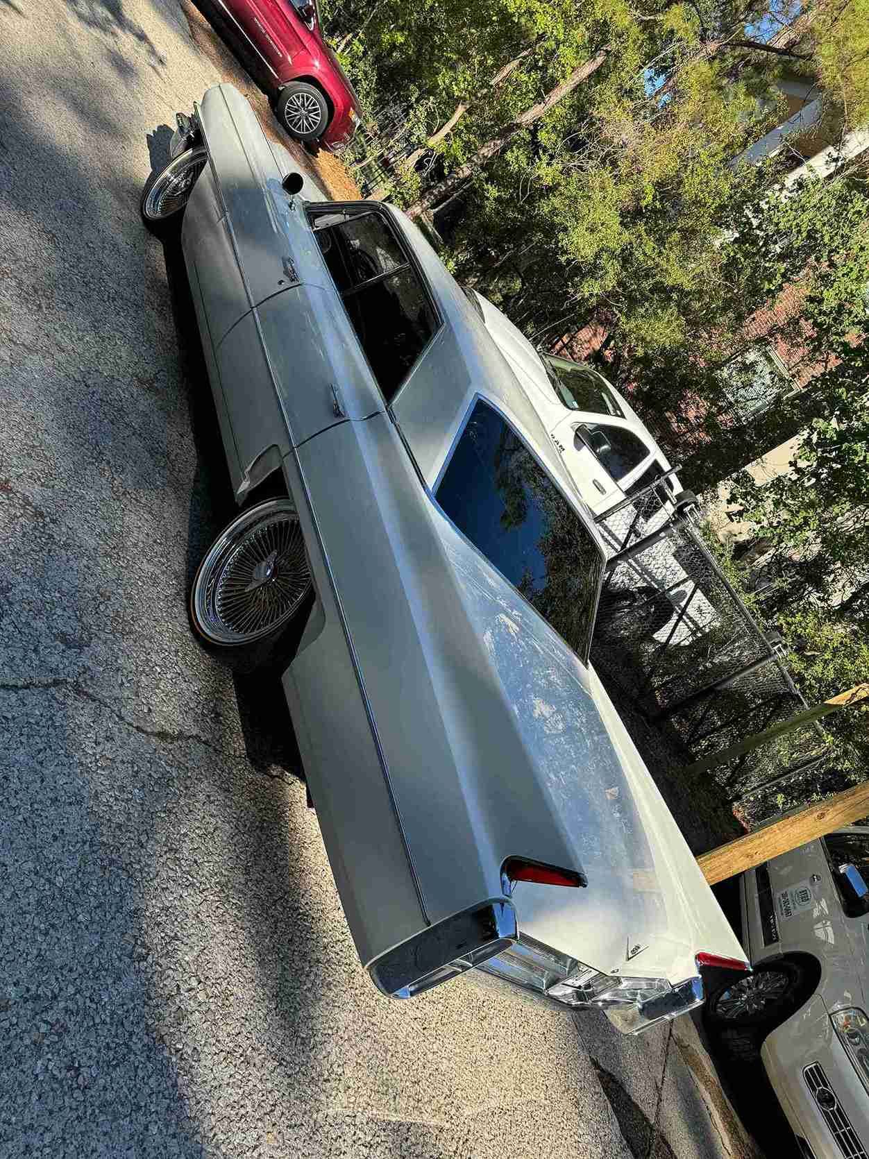 Silver vintage car parked on a driveway with other cars and trees in the background.