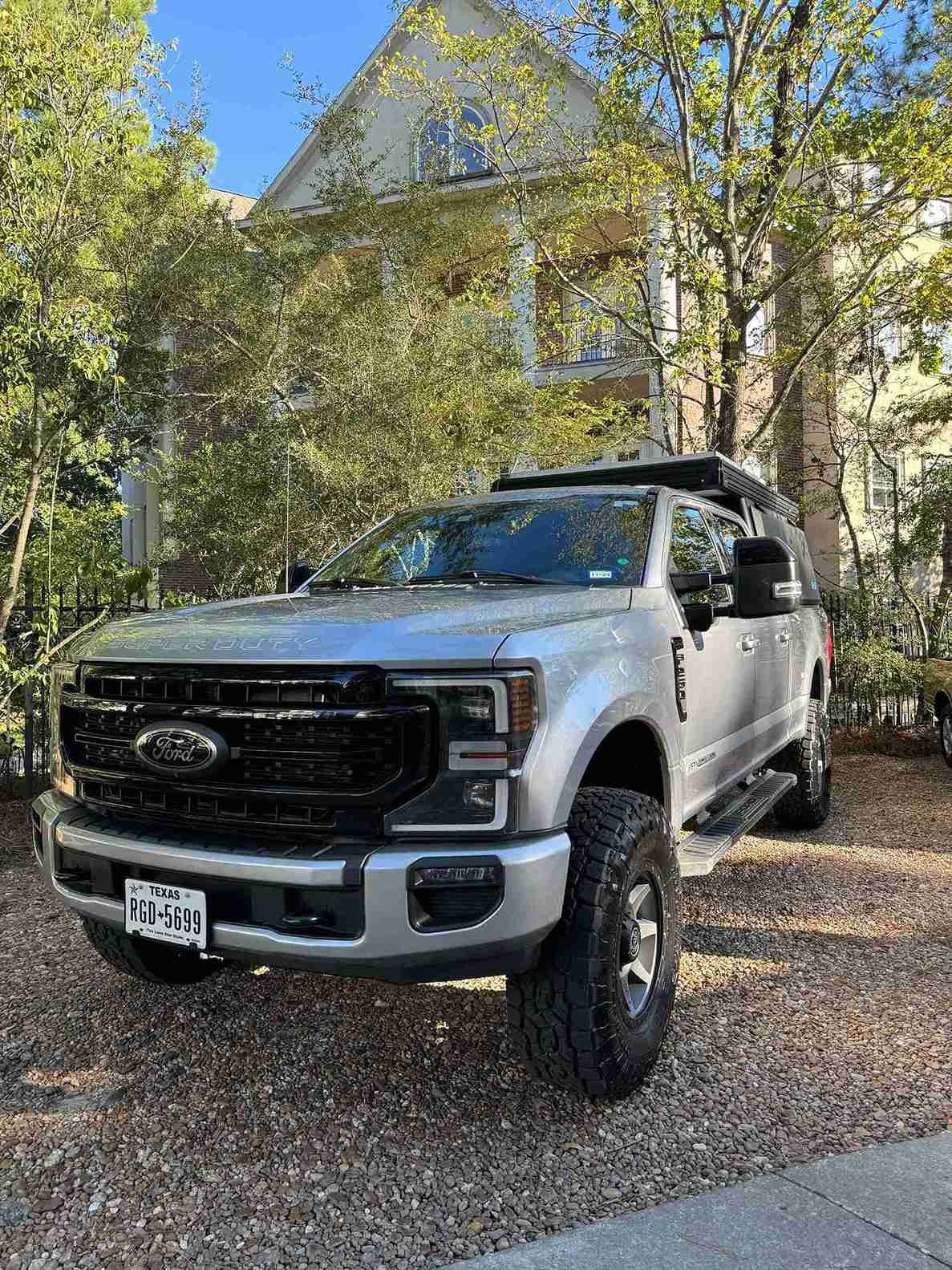 Silver pickup truck parked on a gravel driveway beside a house with trees and a clear blue sky