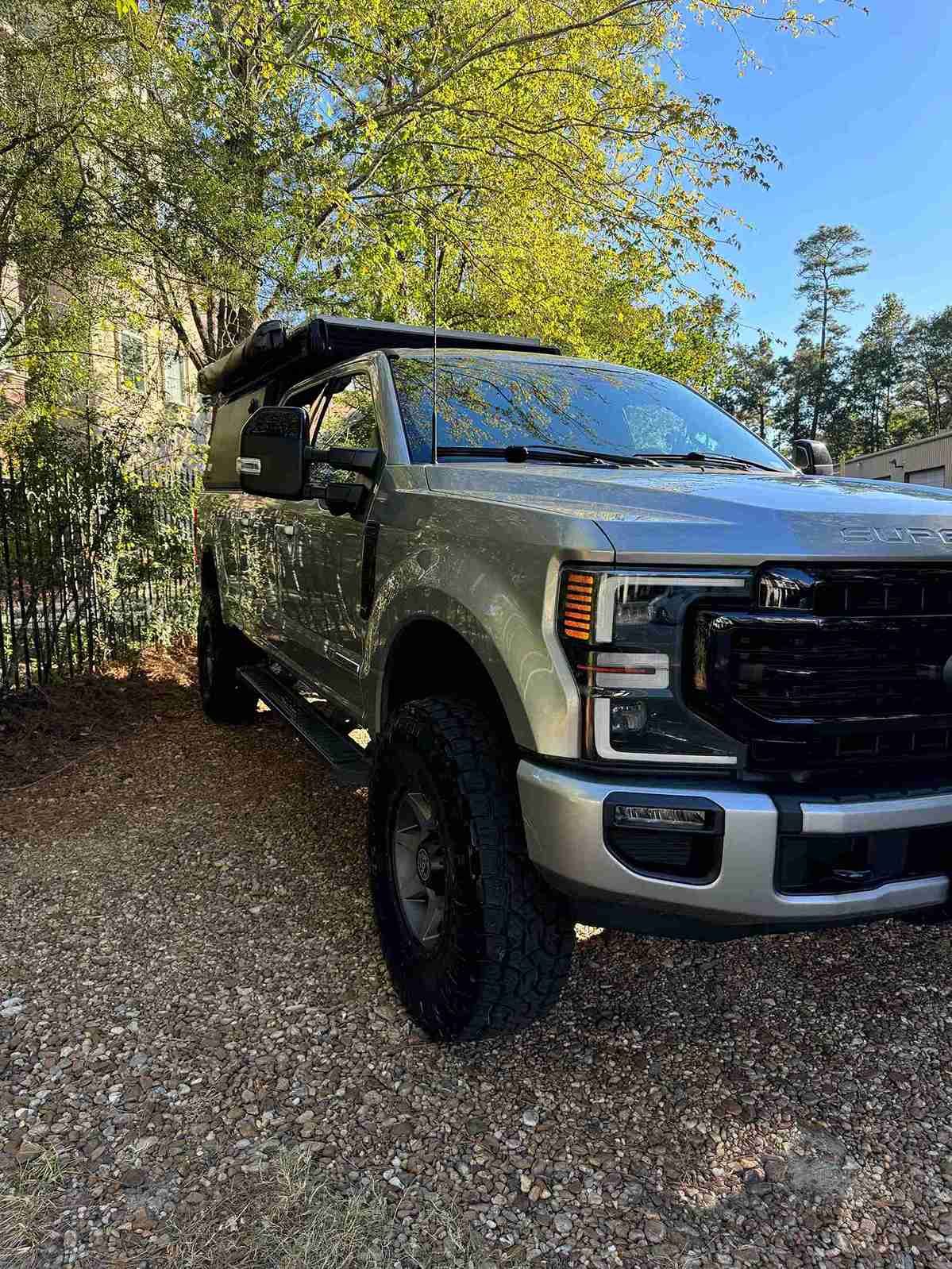 Silver pickup truck with rooftop tent parked on a wooded gravel driveway