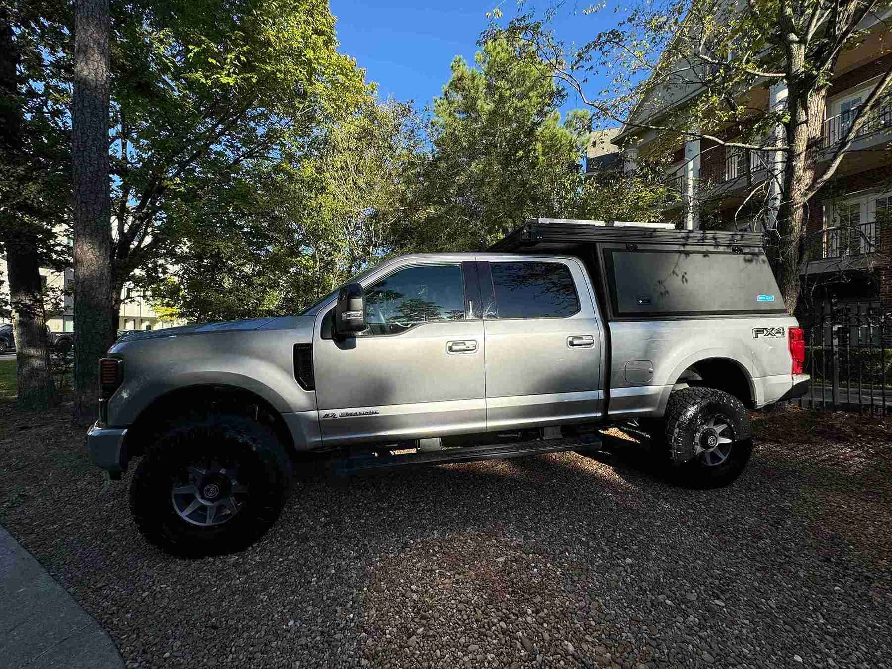 Silver pickup truck parked on a gravel driveway beside trees and a house