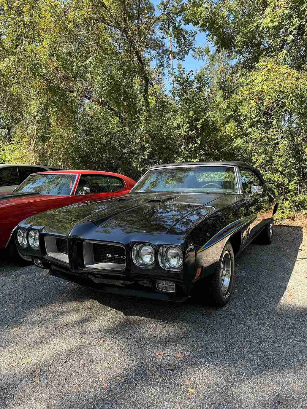 Two classic cars, a black car in front of a red car, parked on gravel under trees