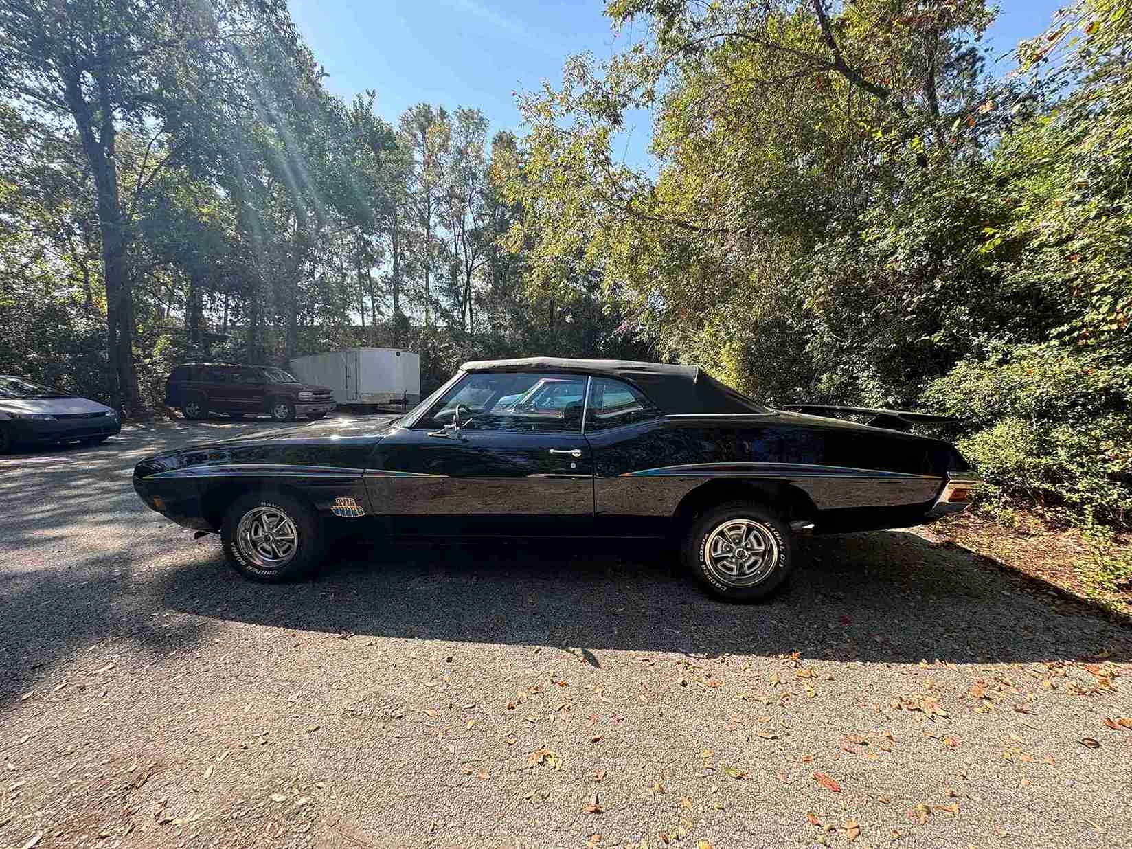 Black classic car parked on a gravel driveway beside trees on a sunny day