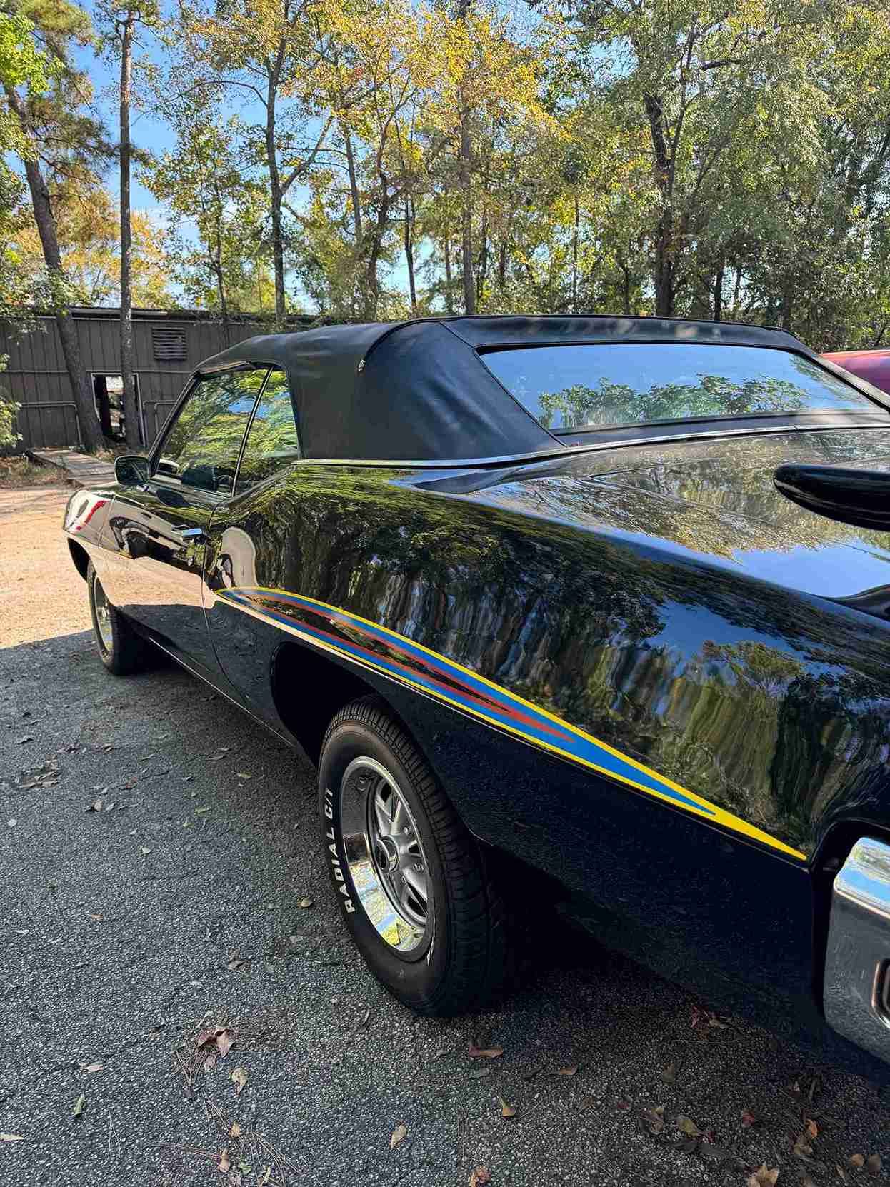 Black classic car with a rainbow stripe parked on gravel near trees, side view