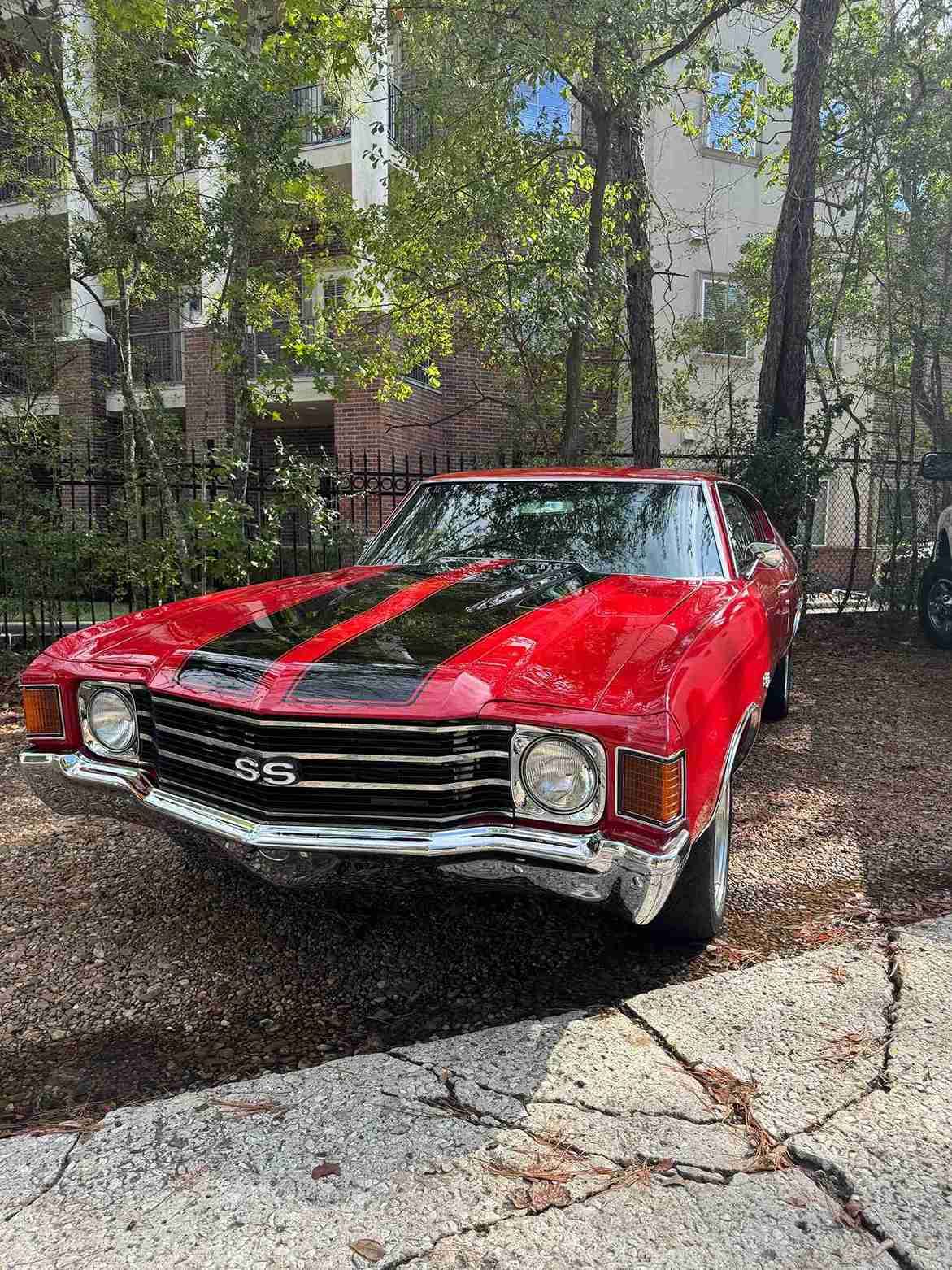 Red classic car with black racing stripes parked outdoors among trees and apartment buildings