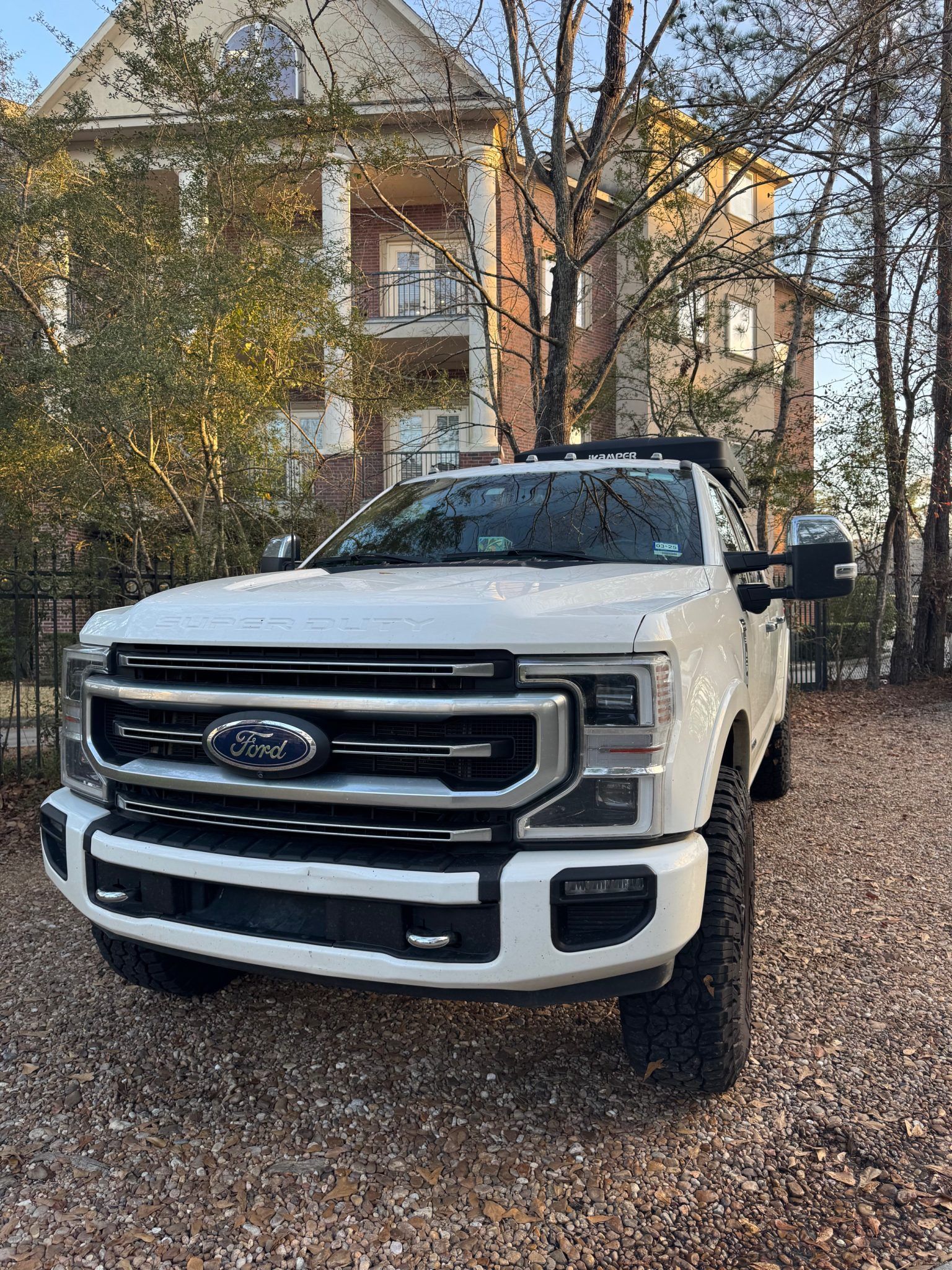 White Ford pickup truck parked on a gravel driveway in front of a house with trees.
