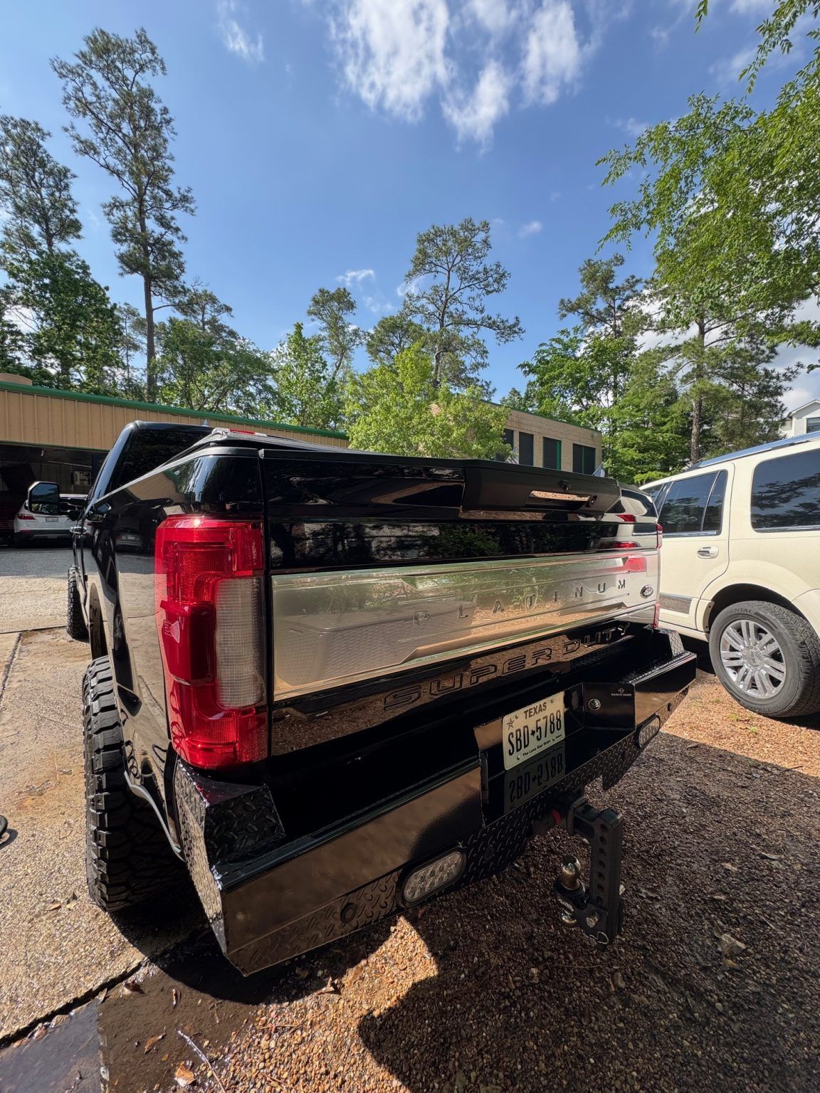 Black pickup truck parked on a driveway beside a white SUV, seen from the rear in a wooded area.