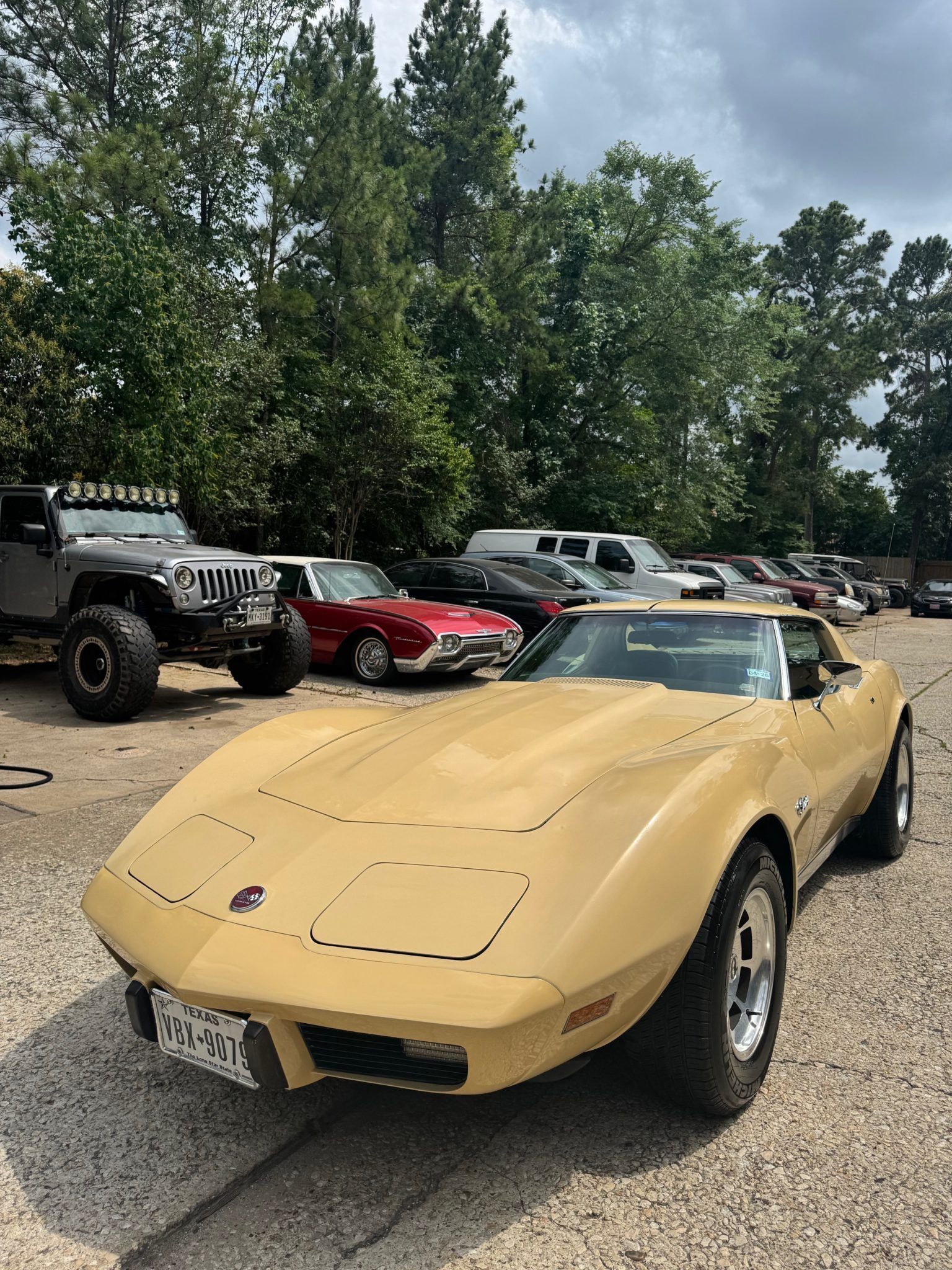 Yellow classic sports car parked on gravel at an outdoor car show, with other cars and trees in the background