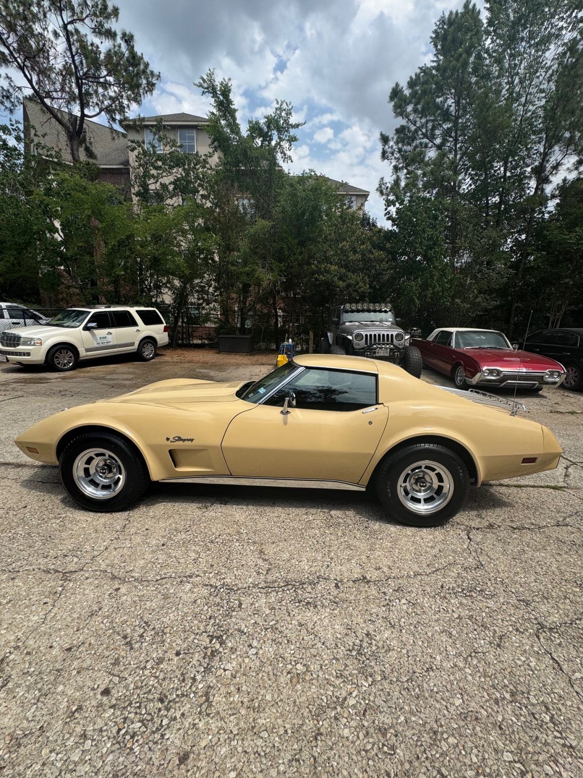 Beige classic sports car parked on a gravel lot with trees and other cars in the background