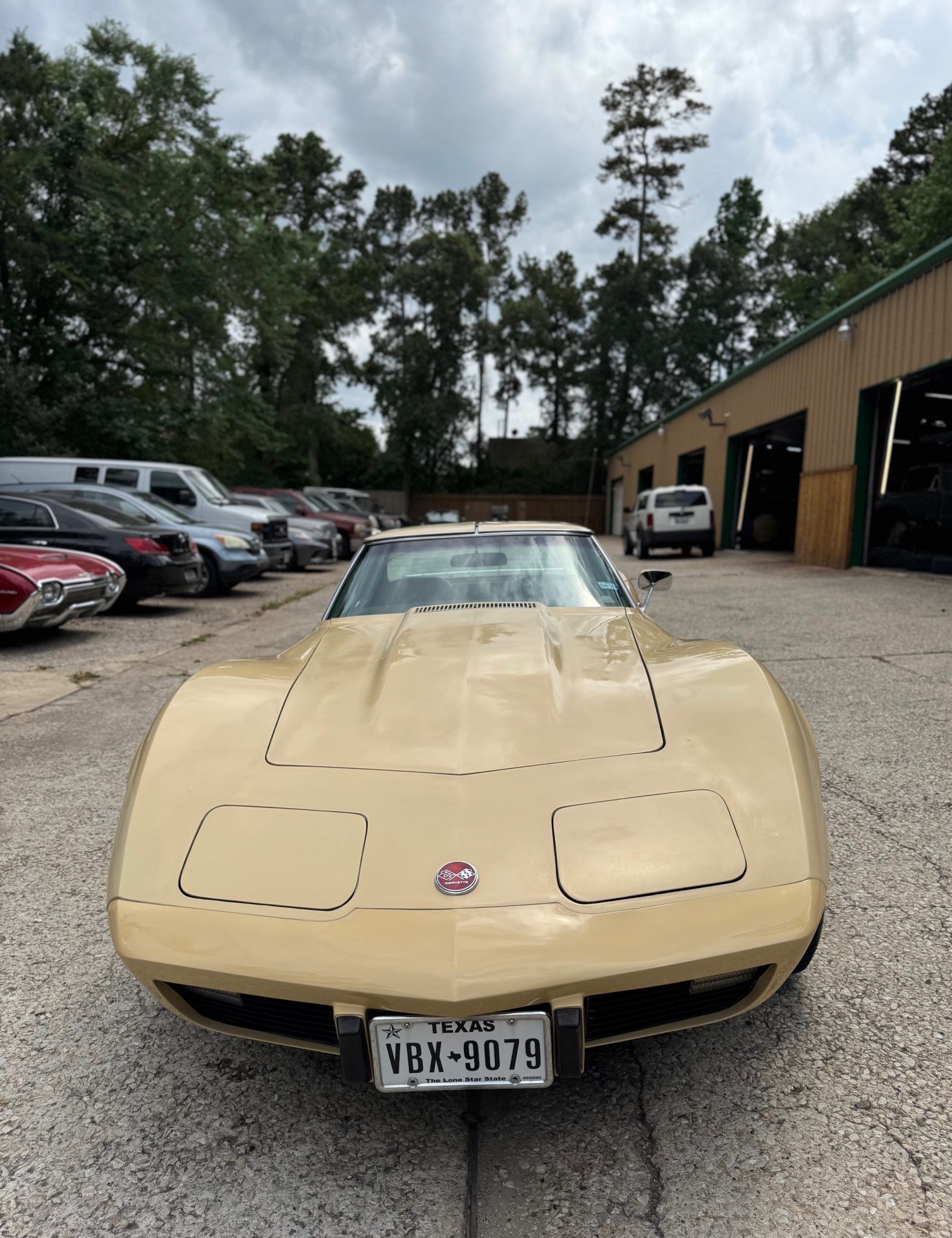 Beige classic car parked on gravel beside a garage and trees