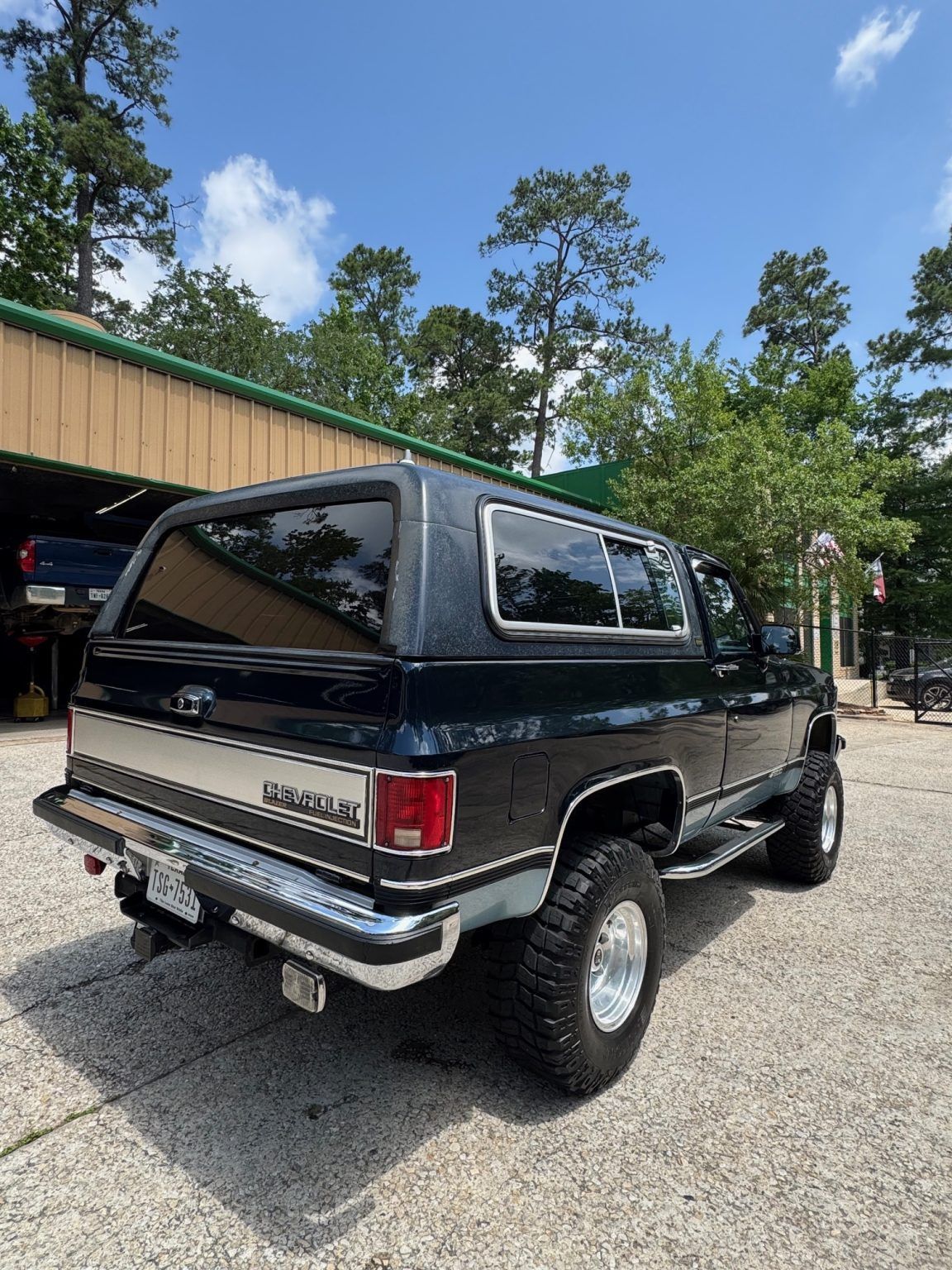 Black lifted SUV parked outdoors beside a garage under trees and blue sky