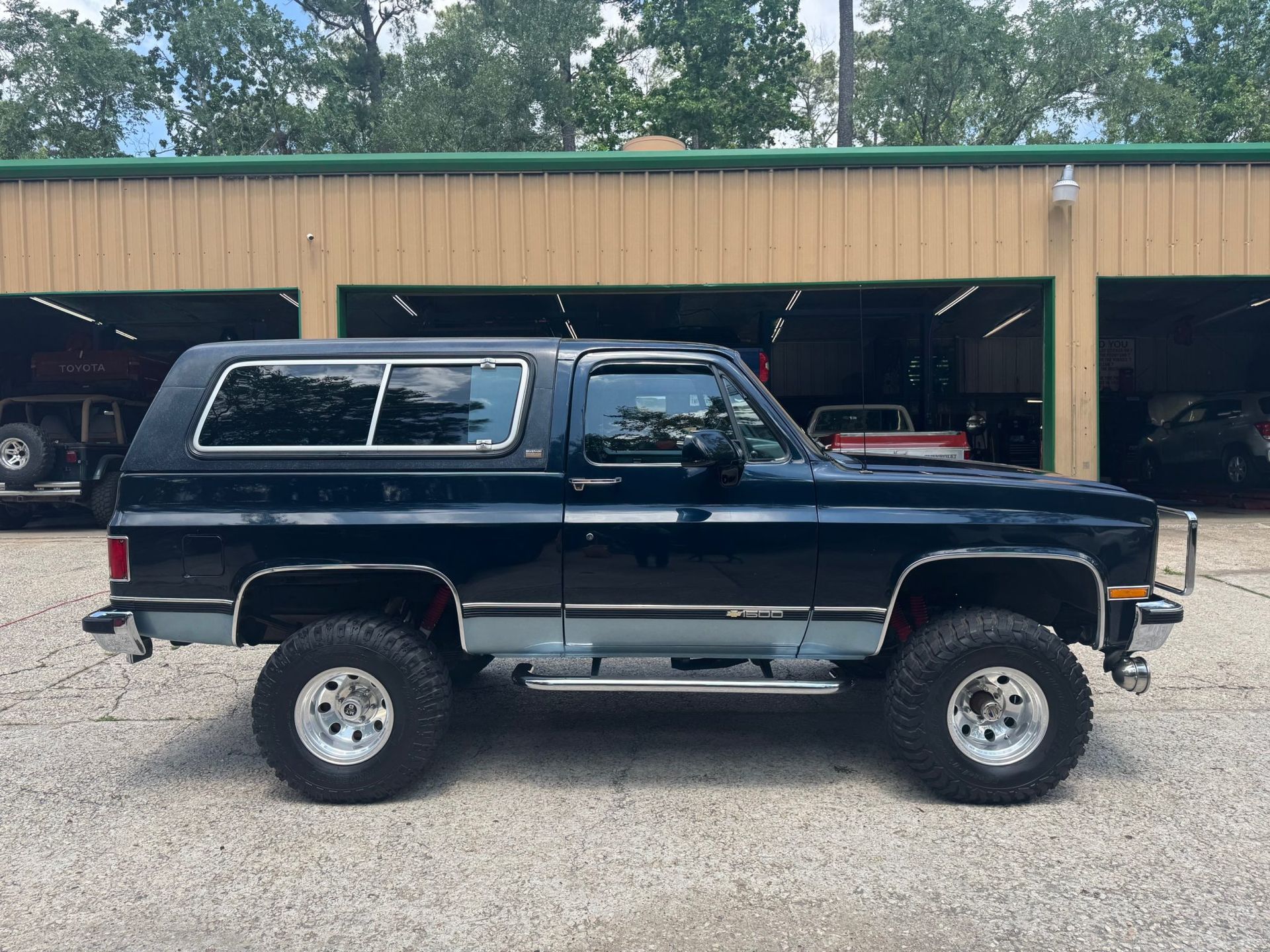 Black lifted SUV with camper shell parked on gravel in front of an open garage