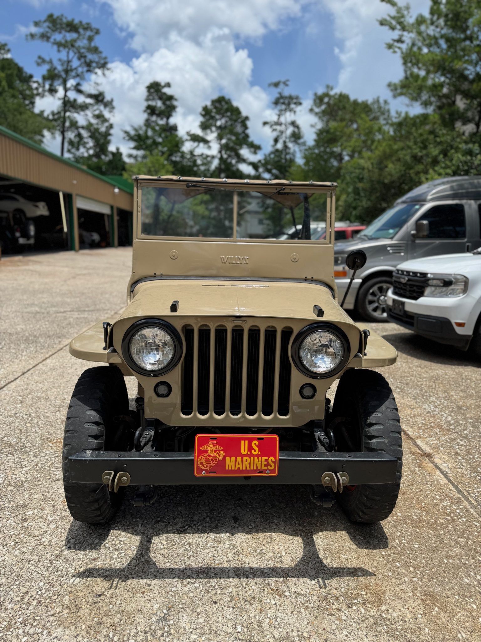 Front view of a tan vintage Jeep parked outdoors on a gravel lot beside other vehicles.