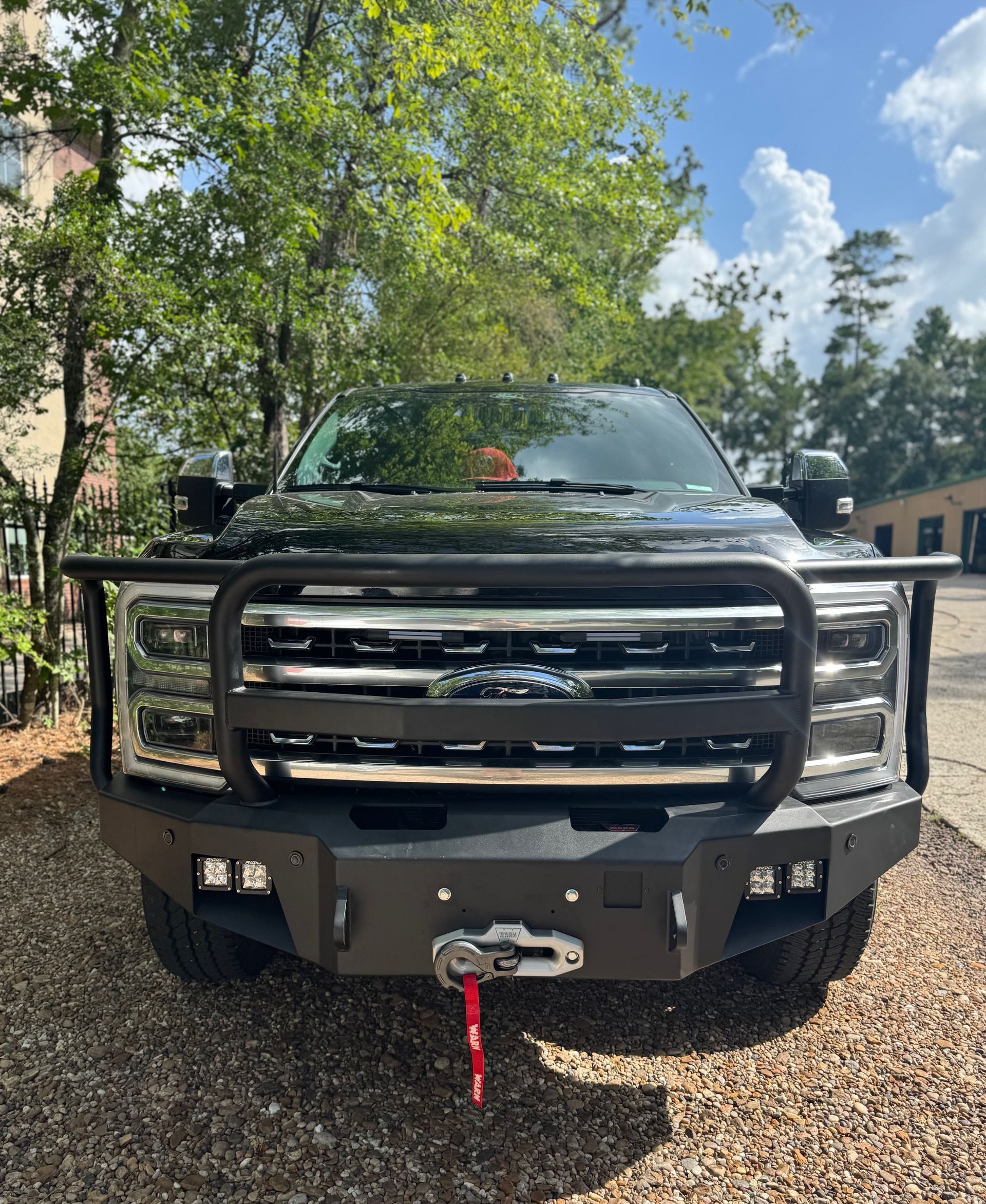 Black pickup truck with a heavy front grille parked on gravel under trees, front view