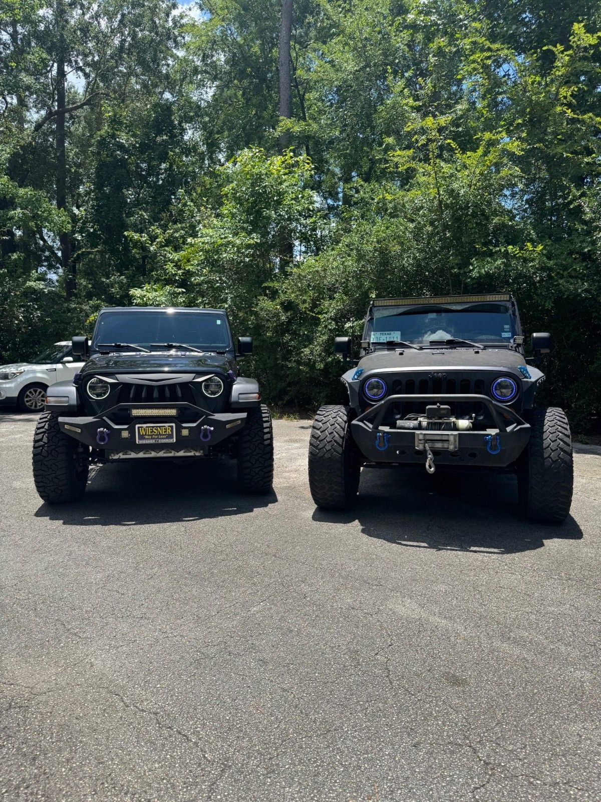 Two black Jeeps parked side by side in a sunny lot with trees in the background