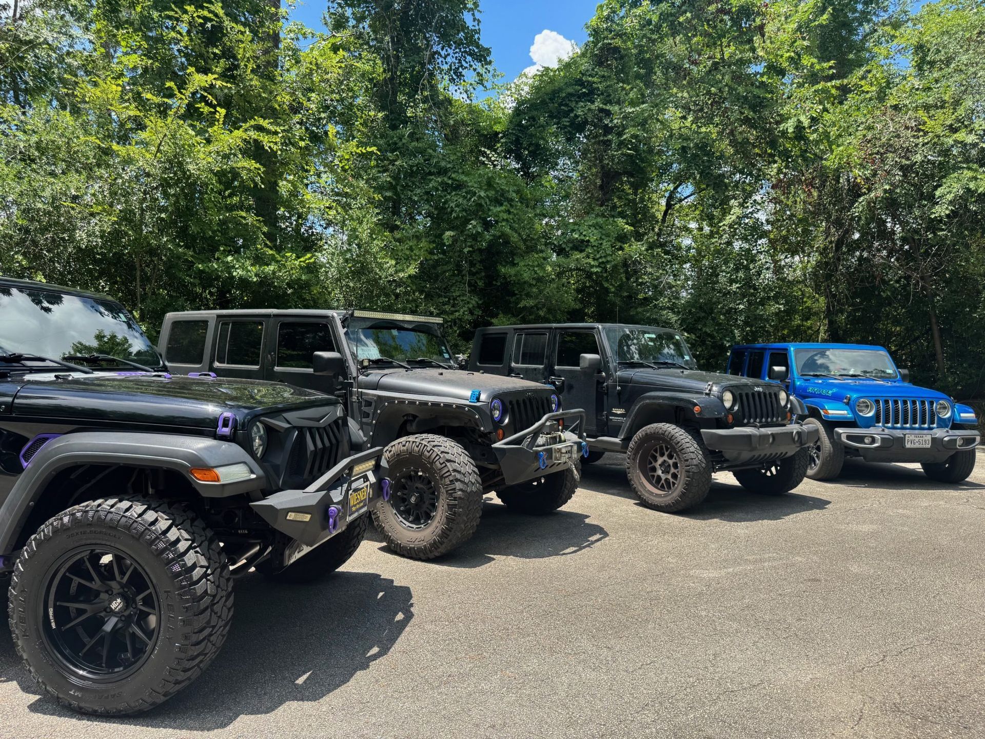 Three black and blue Jeeps parked on a gravel lot under trees and a bright sky