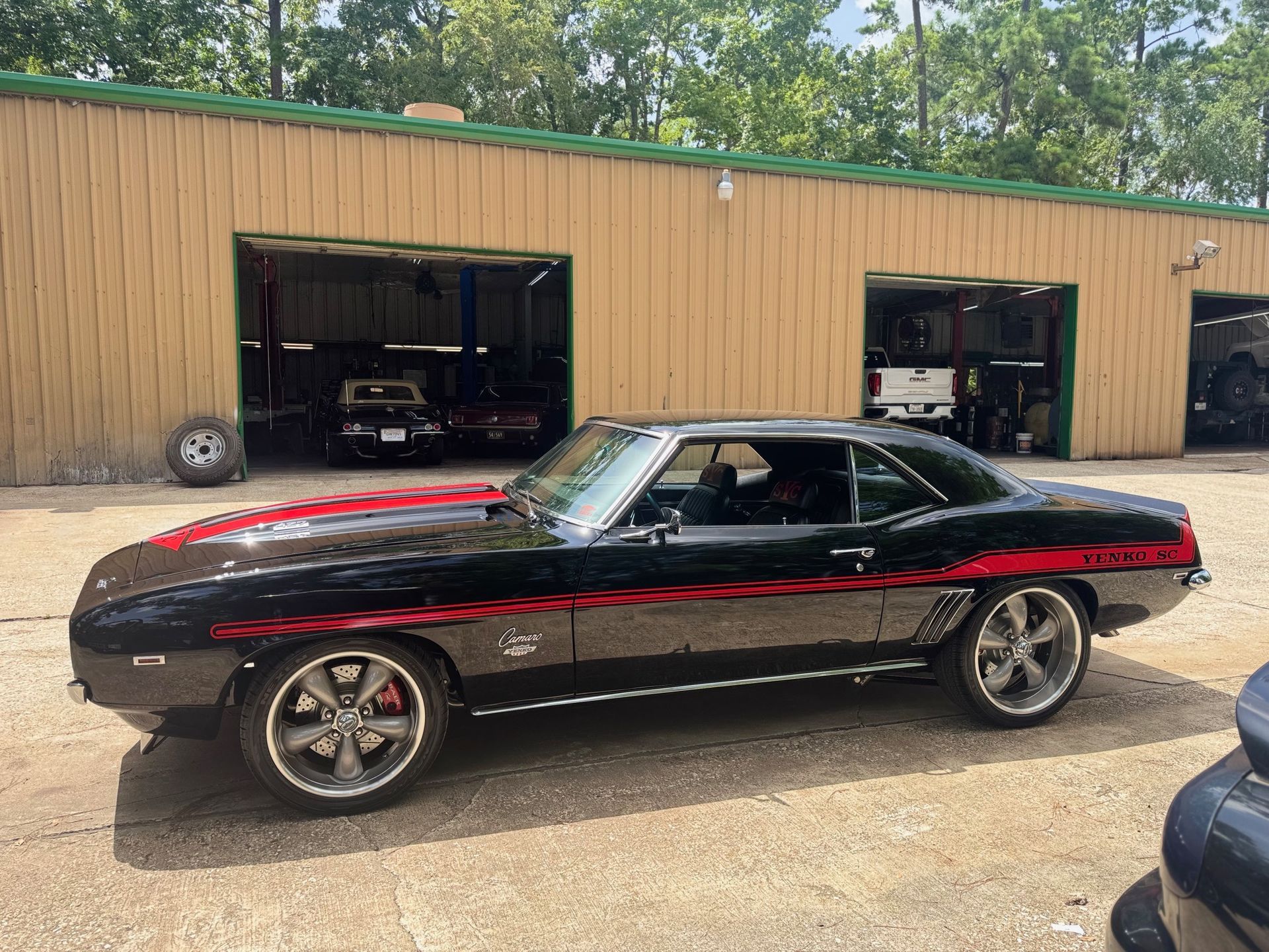 Black classic muscle car with red stripe parked outside an auto shop garage.