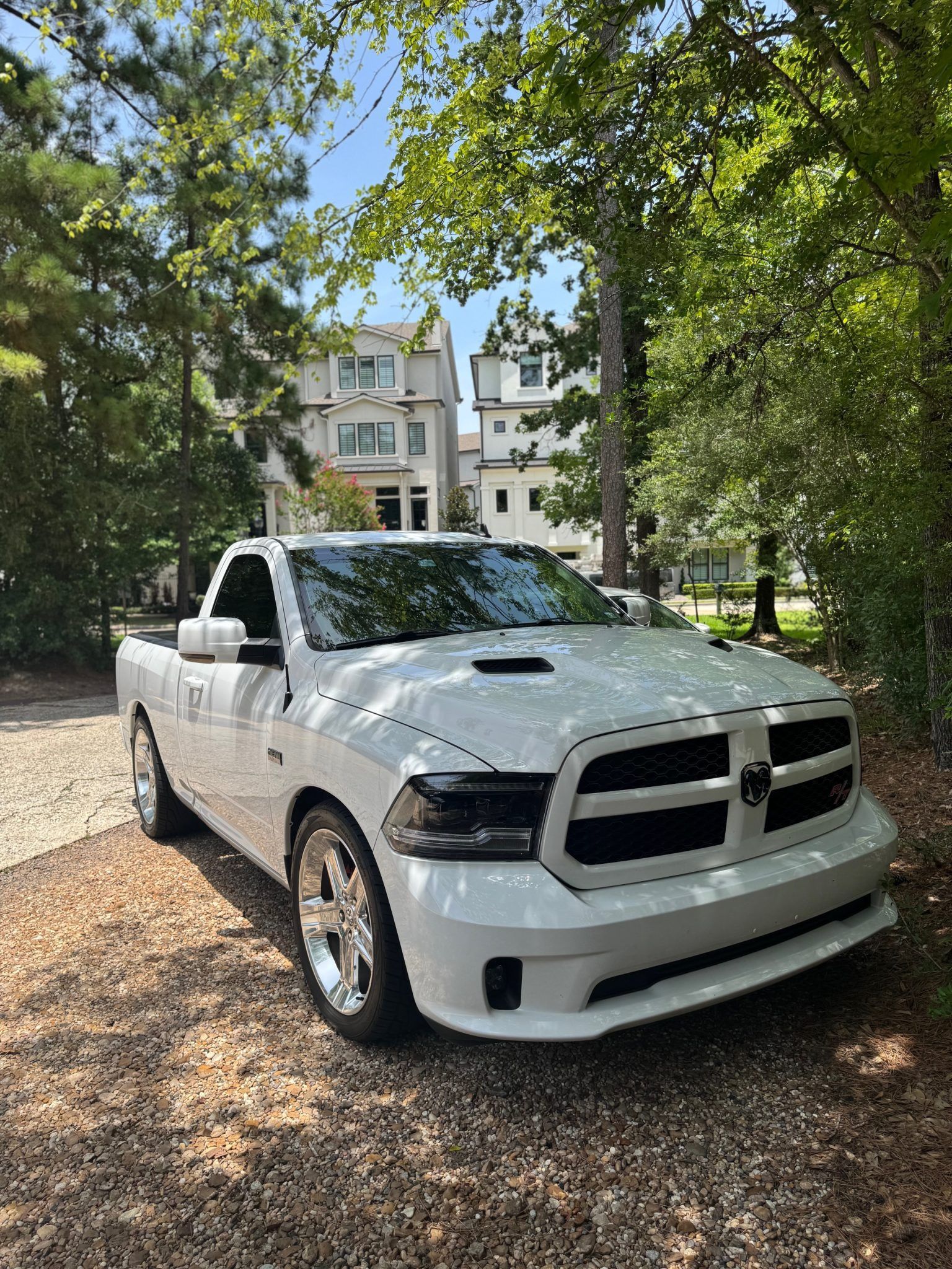 White pickup truck parked on a leaf-covered driveway beneath trees near a residential building.