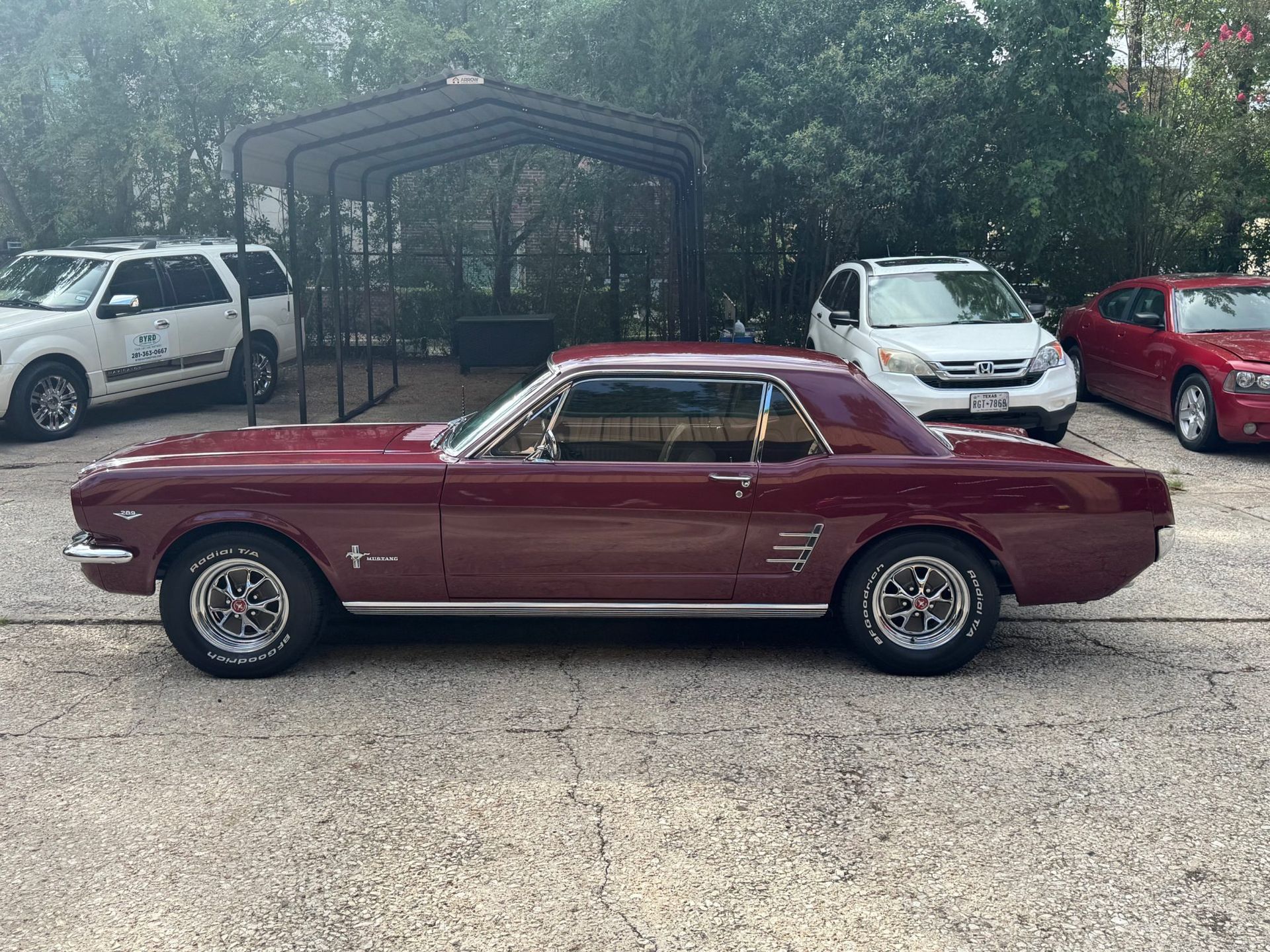 Maroon classic coupe parked in a lot, with other cars and a covered structure in the background